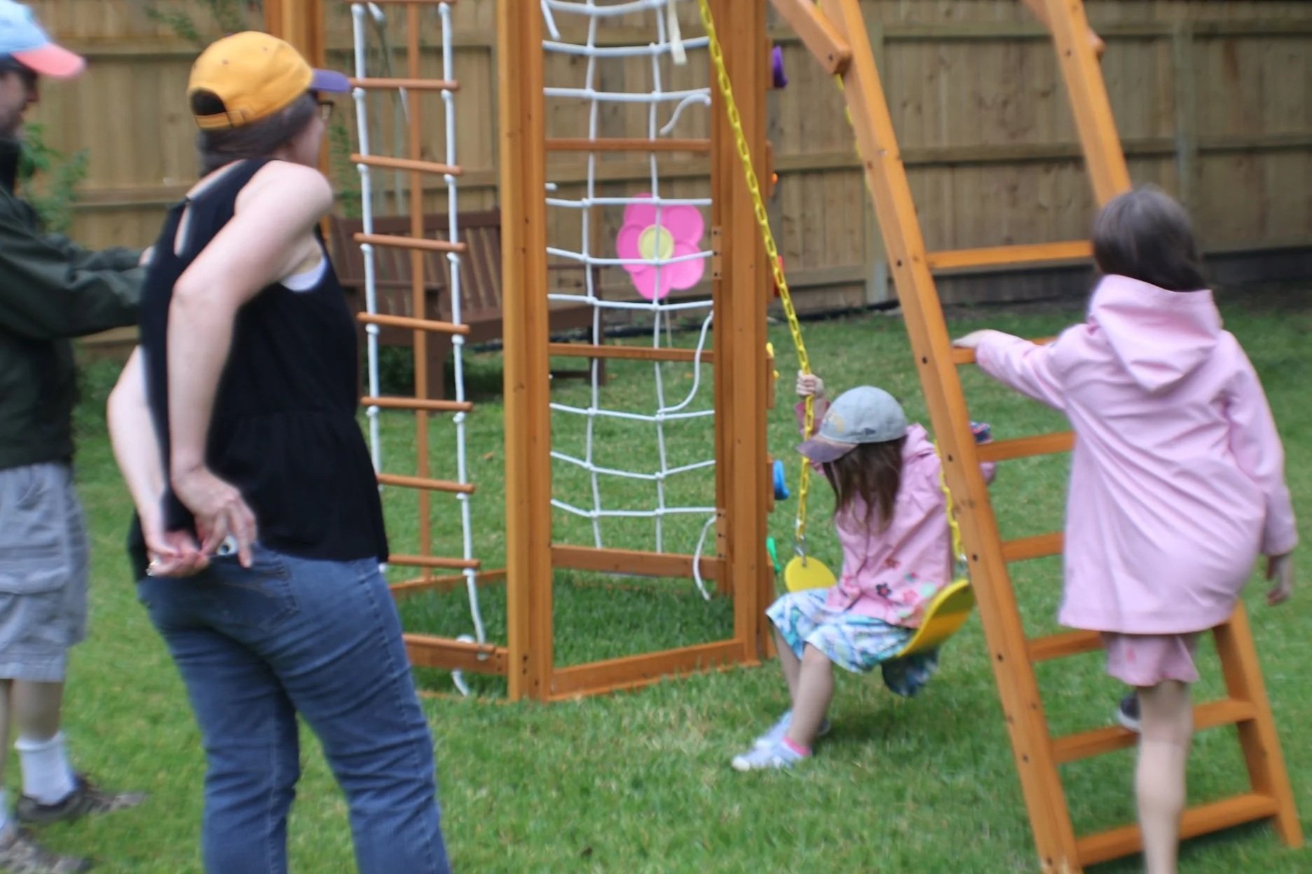 A backyard scene with two children playing on a wooden swing set and others standing nearby. A woman is observing. The scene includes grass and a wooden fence in the background.