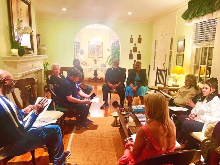 A group of people sitting in a living room arranged for a meeting, with some taking notes, and a person at the head of the room speaking.