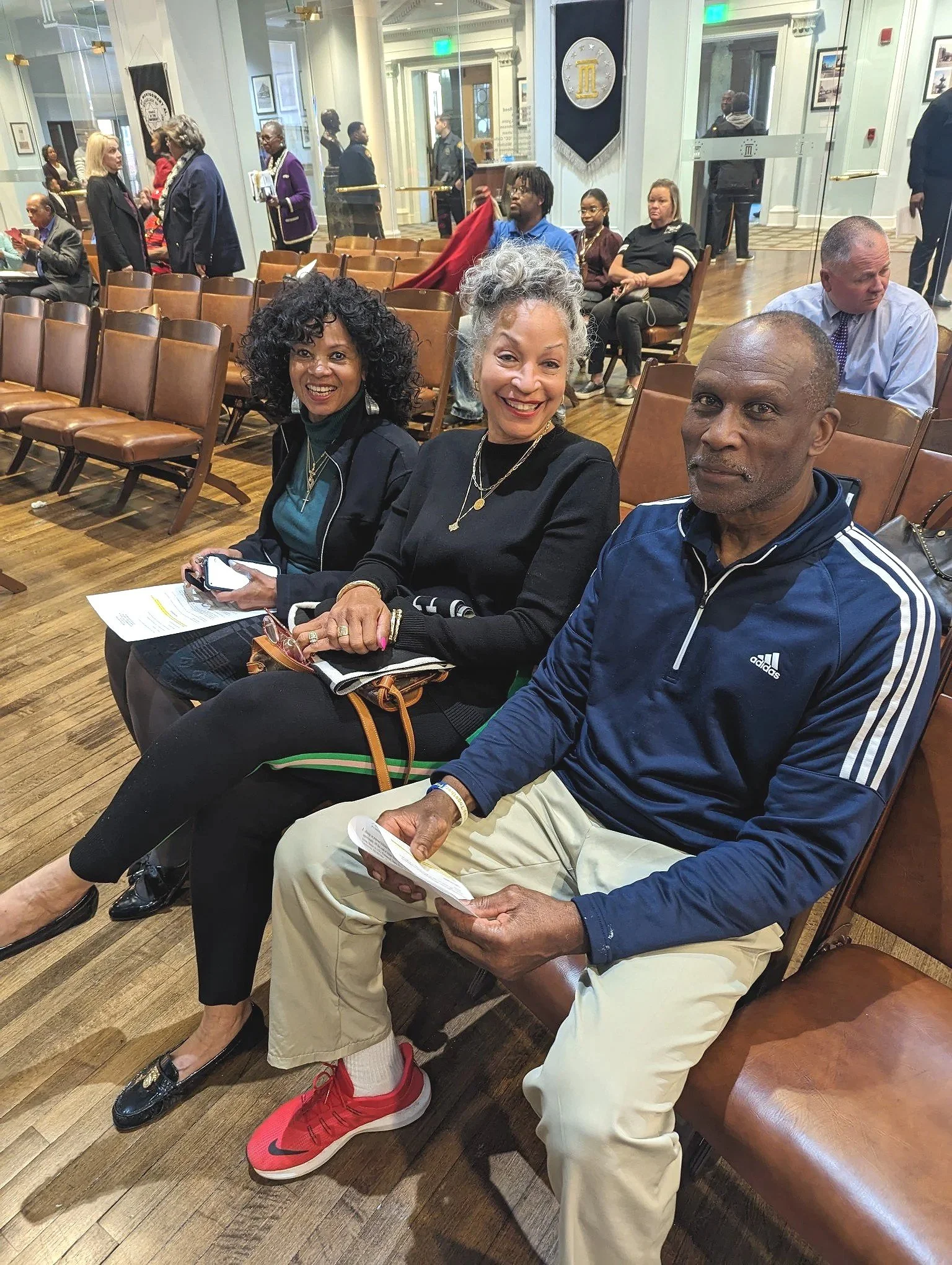 Three people sitting in a row of chairs inside a room, smiling at the camera. The woman in the middle has gray curly hair, the man on the right has a shaved head, and the woman on the left has curly black hair. They are holding papers and sitting next to each other.