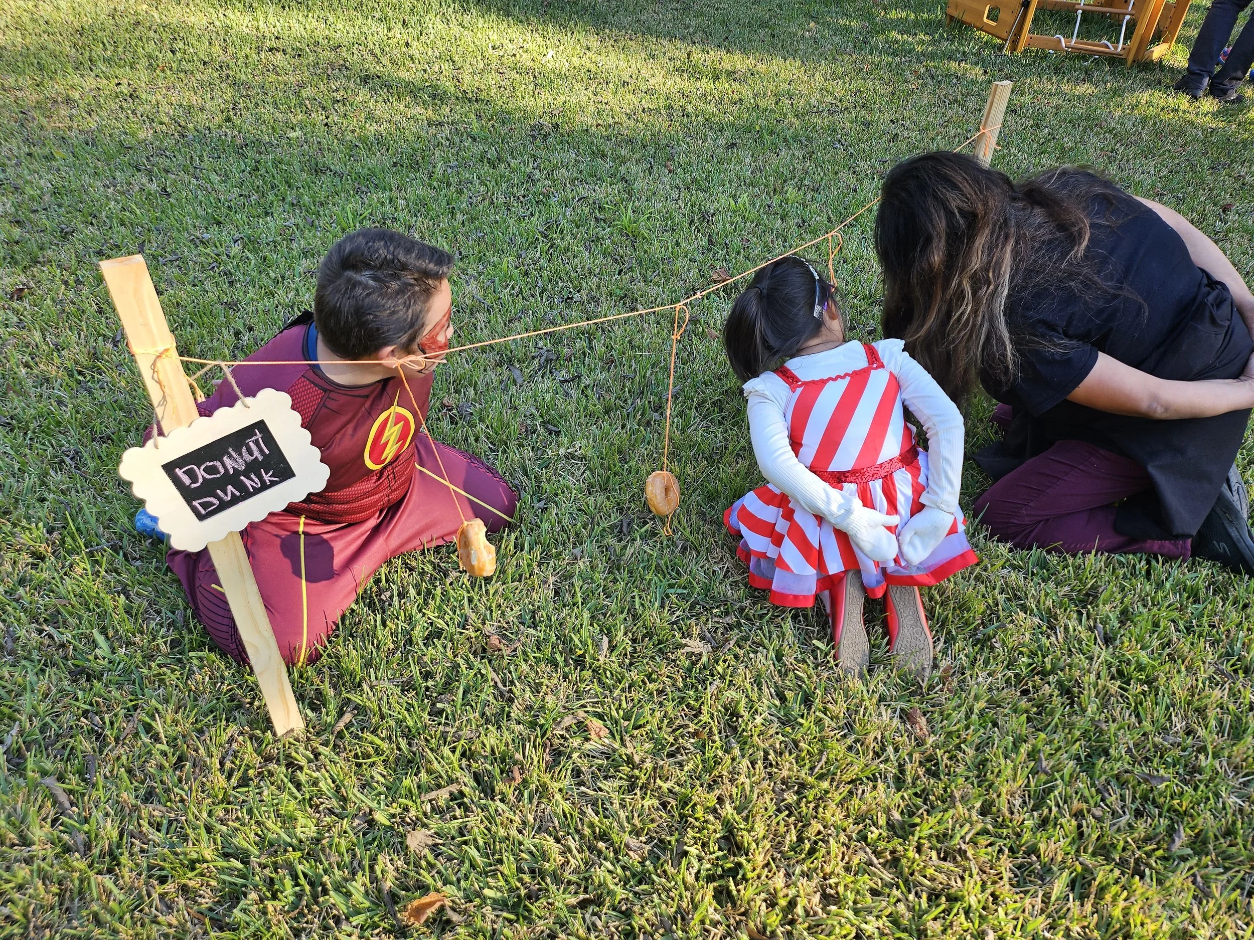 Children and an adult participating in a pumpkin dunking game outdoors on grass, with a small chalkboard sign that reads 'Dunk It Dunk' and a pumpkin hanging from a string.