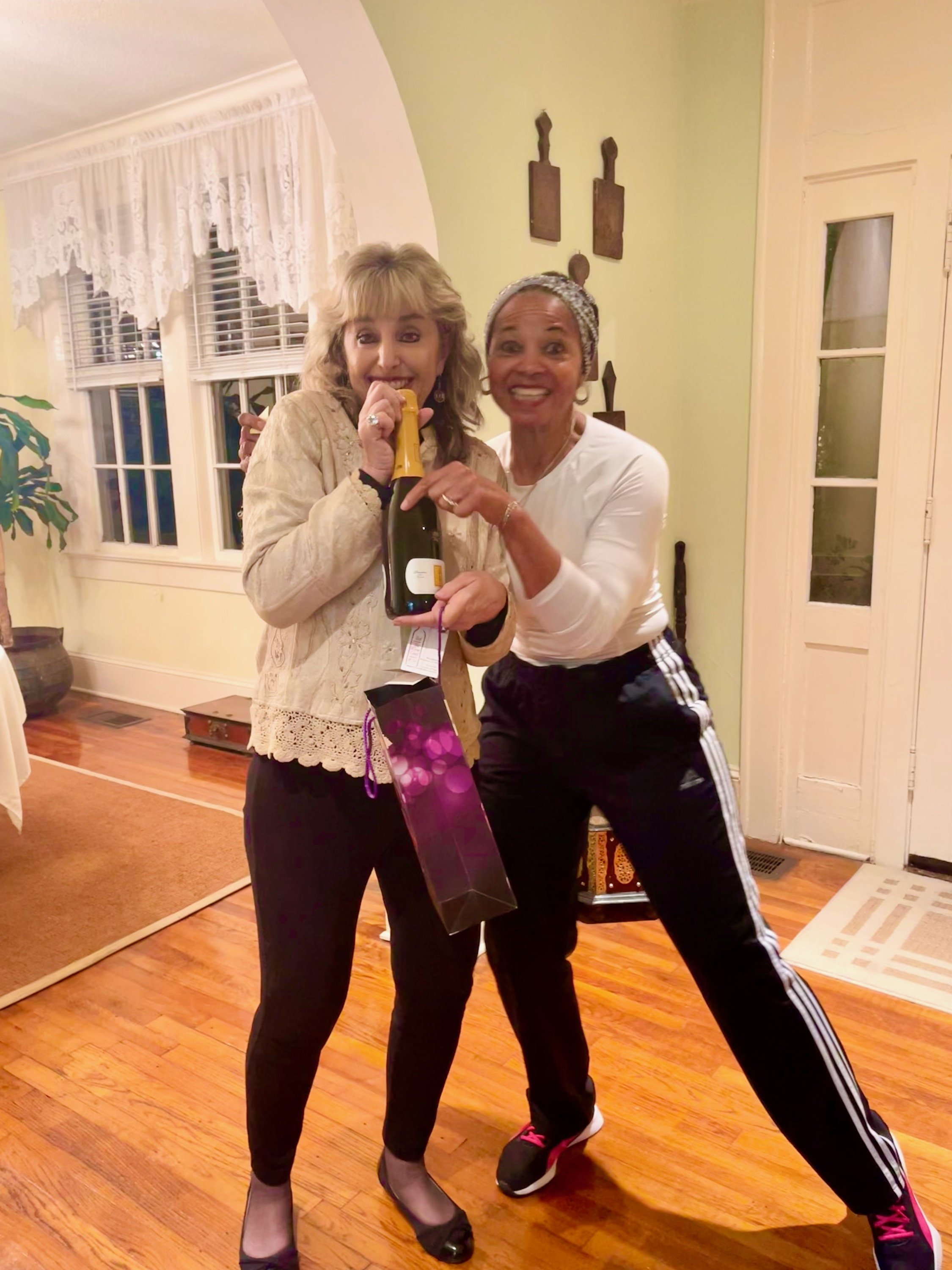 Two women smiling and dancing indoors, holding a bottle of sparkling wine, with a background of windows with lace curtains and wooden decorations on the wall.