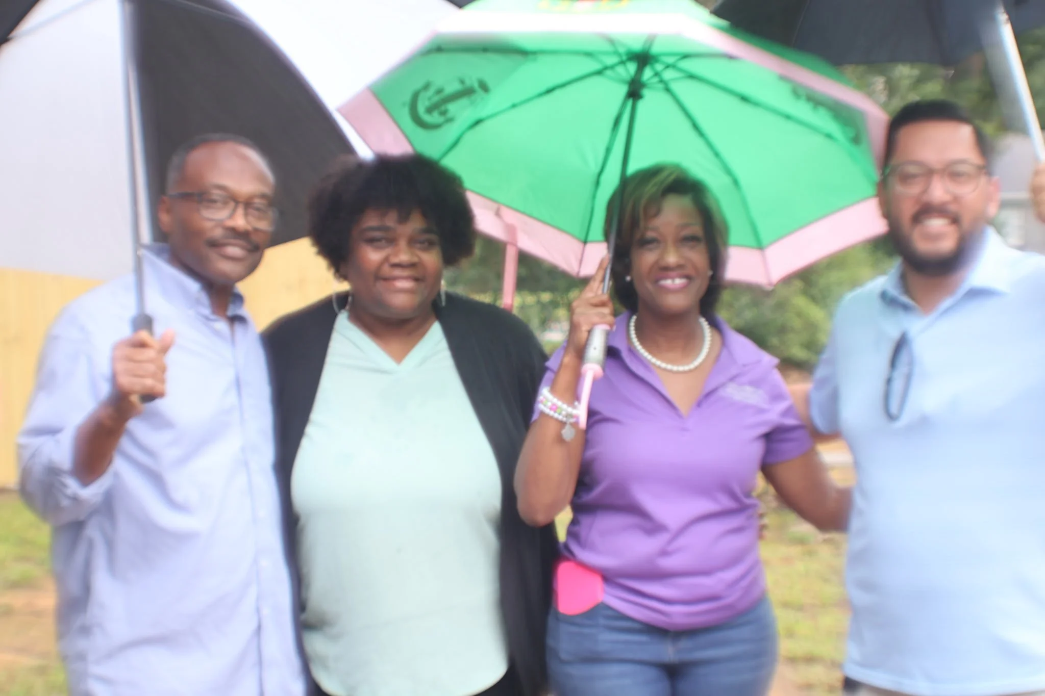 Group of four people standing outdoors under a green and pink umbrella, smiling at the camera.