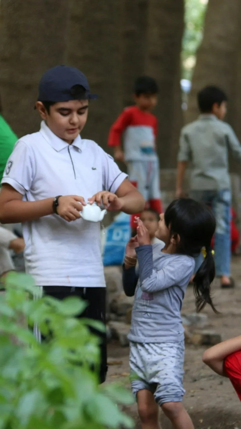 A young boy in a white shirt and navy cap is pouring water into a girl's hands in a natural outdoor setting with trees. Other children are in the background.