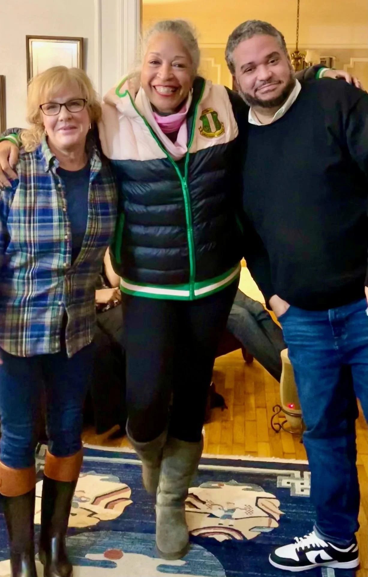 Three people standing together indoors, smiling, with a woman in the middle. The woman in the middle is wearing a black puffer vest with a crest on it, gray pants, and gray boots. The woman on the left is wearing glasses, a blue shirt, a plaid jacket, and brown boots. The man on the right is wearing a black sweater, white shirt, jeans, and black-and-white sneakers.