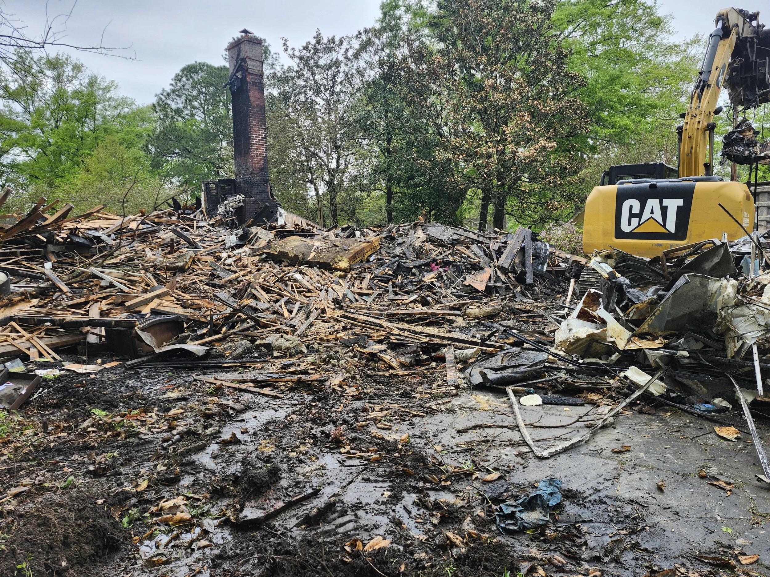 Scene of a house fire with a burnt chimney, debris, and destroyed remains of a house, with a yellow Caterpillar excavator on the right side, surrounded by trees.