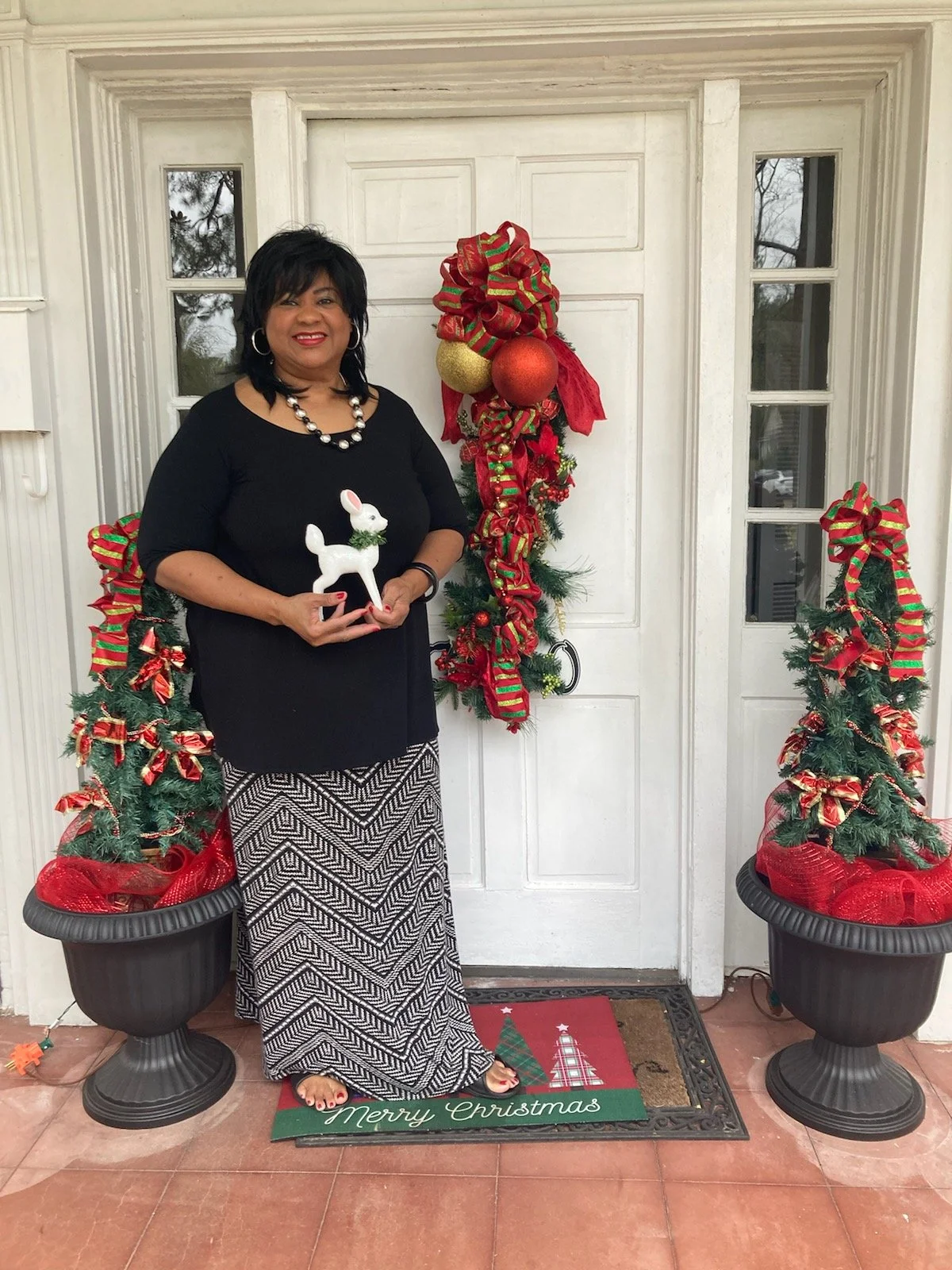 Woman standing in front of a decorated Christmas door, holding a white reindeer ornament, with Christmas wreaths and trees on either side.
