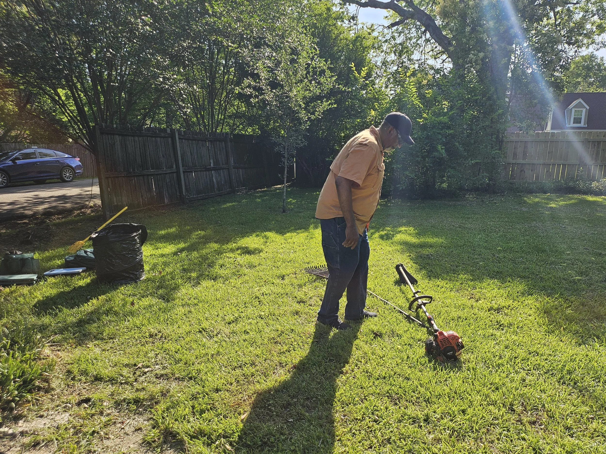 A man wearing a tan shirt, jeans, sunglasses and a cap is working outdoors with a gas-powered tool on a sunny day. There are bags, tools, and gardening equipment on the grass nearby, and a wooden fence, trees, and a house are in the background.