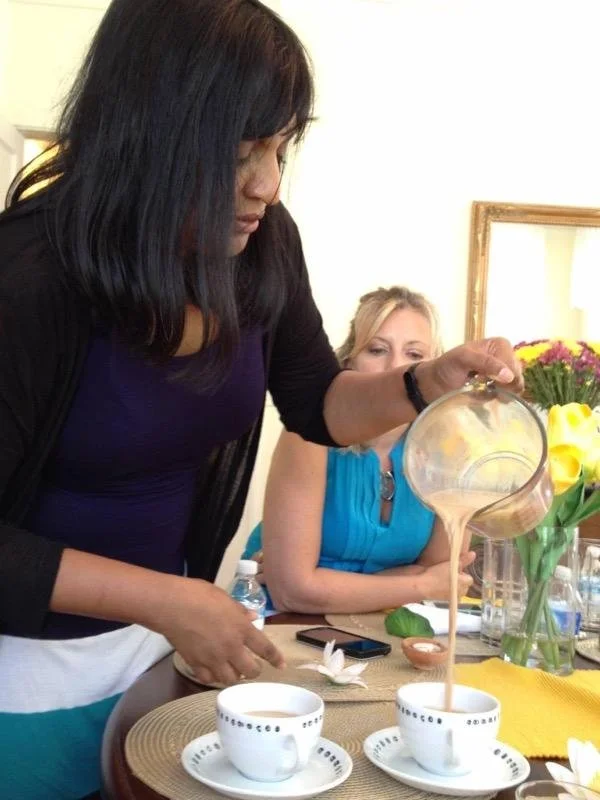 Woman pouring beige liquid from a pitcher into a teacup on a table with flowers and other cups.