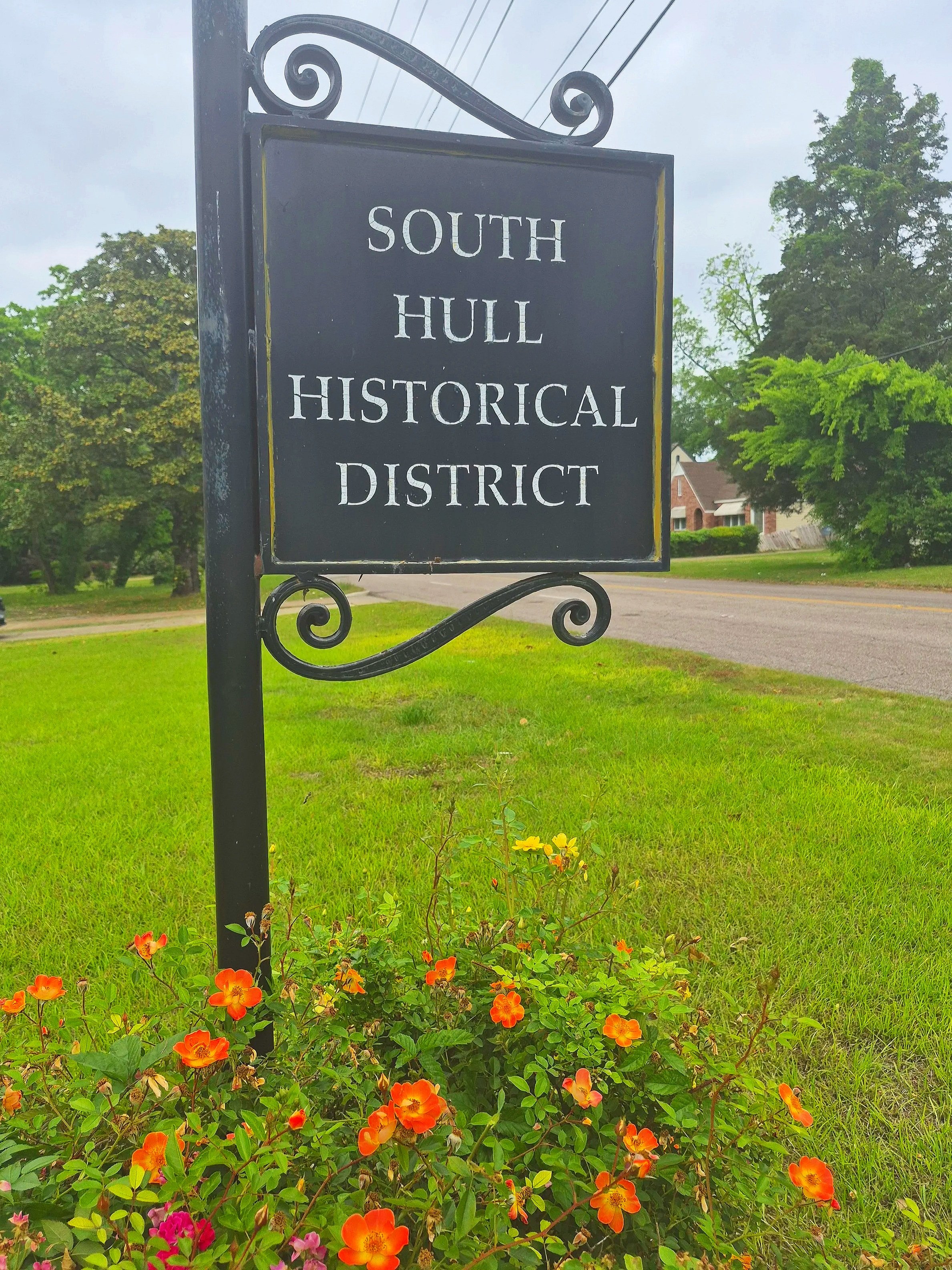 Black sign with white text reading "South Hull Historical District" surrounded by green grass, orange flowers, and trees in the background on a cloudy day.