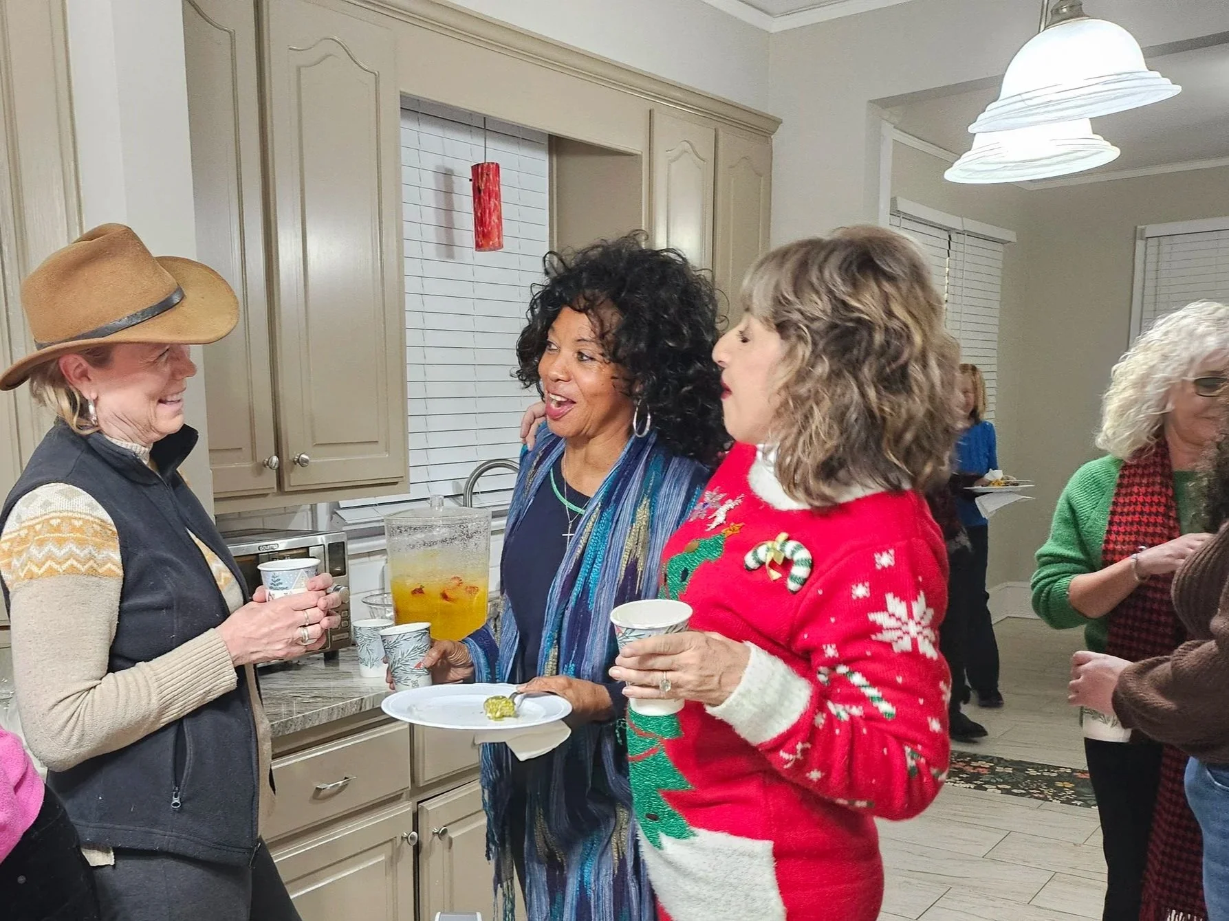 People gathered in a kitchen during a holiday celebration, wearing festive clothing and holding drinks and snacks.