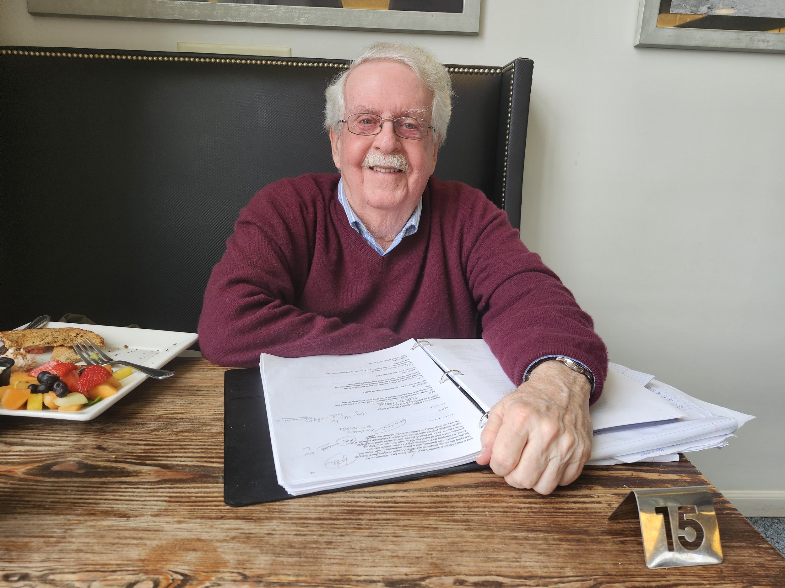 An elderly man with white hair, glasses, and a mustache is sitting at a wooden table, smiling, with an open binder and a pile of papers in front of him. There are plates with fruit and a slice of pie on the table, and a metal table number '15' is visible. The background includes a wall and framed mirrors.