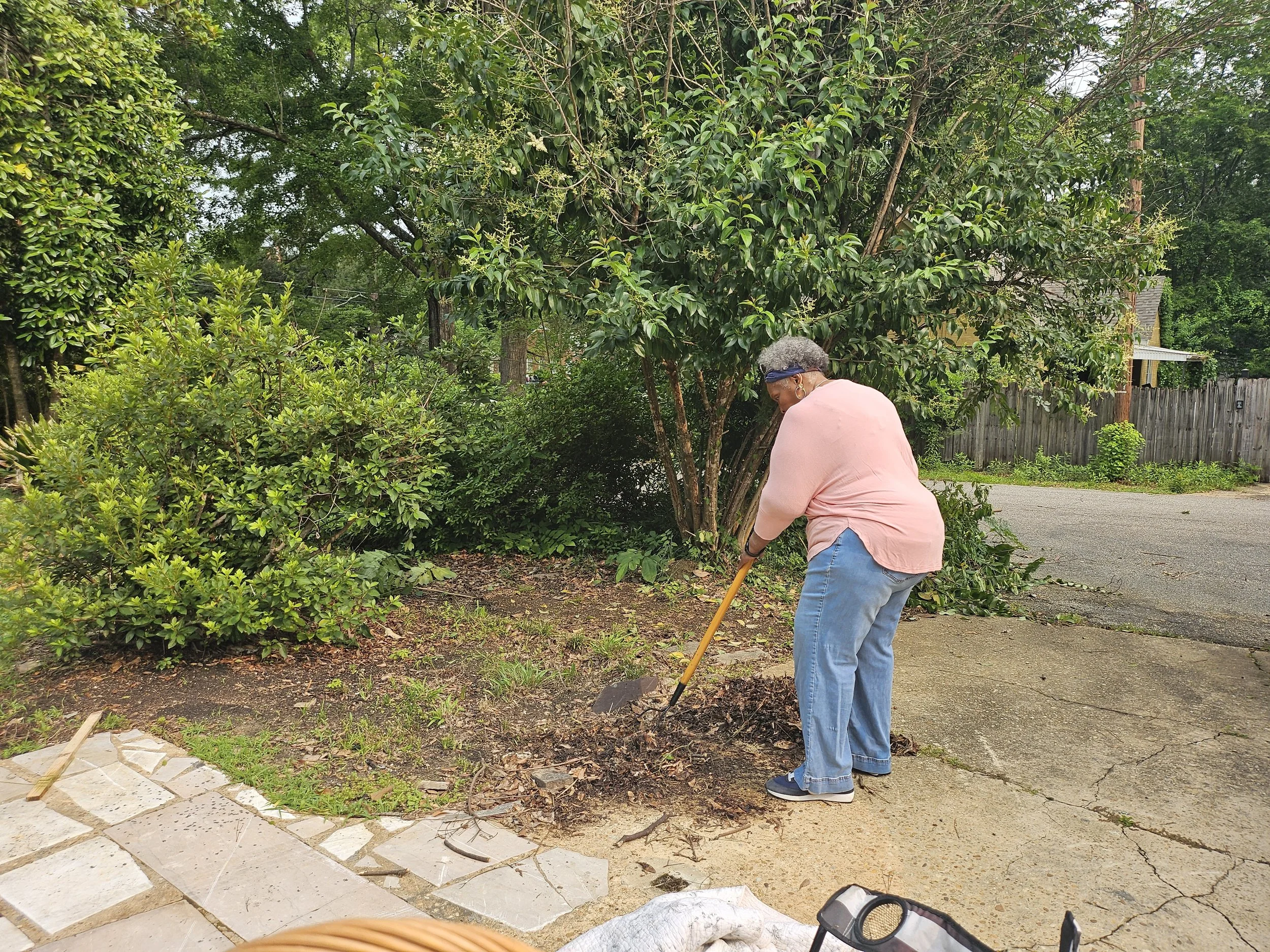 A woman wearing a pink shirt, blue jeans, sneakers, a headband, and earrings is gardening outdoors, using a small gardening tool to tend to plants near a tree and a landscaped area. There is a backyard fence, trees, and garden elements visible in the background.