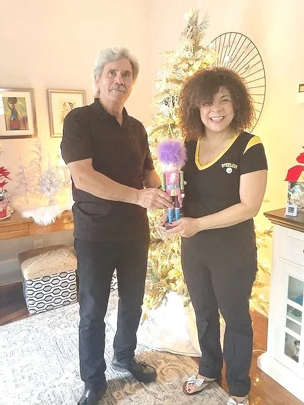 A man and woman standing indoors near a decorated Christmas tree, holding a nutcracker figurine together, smiling, with framed artworks and holiday decorations in the background.