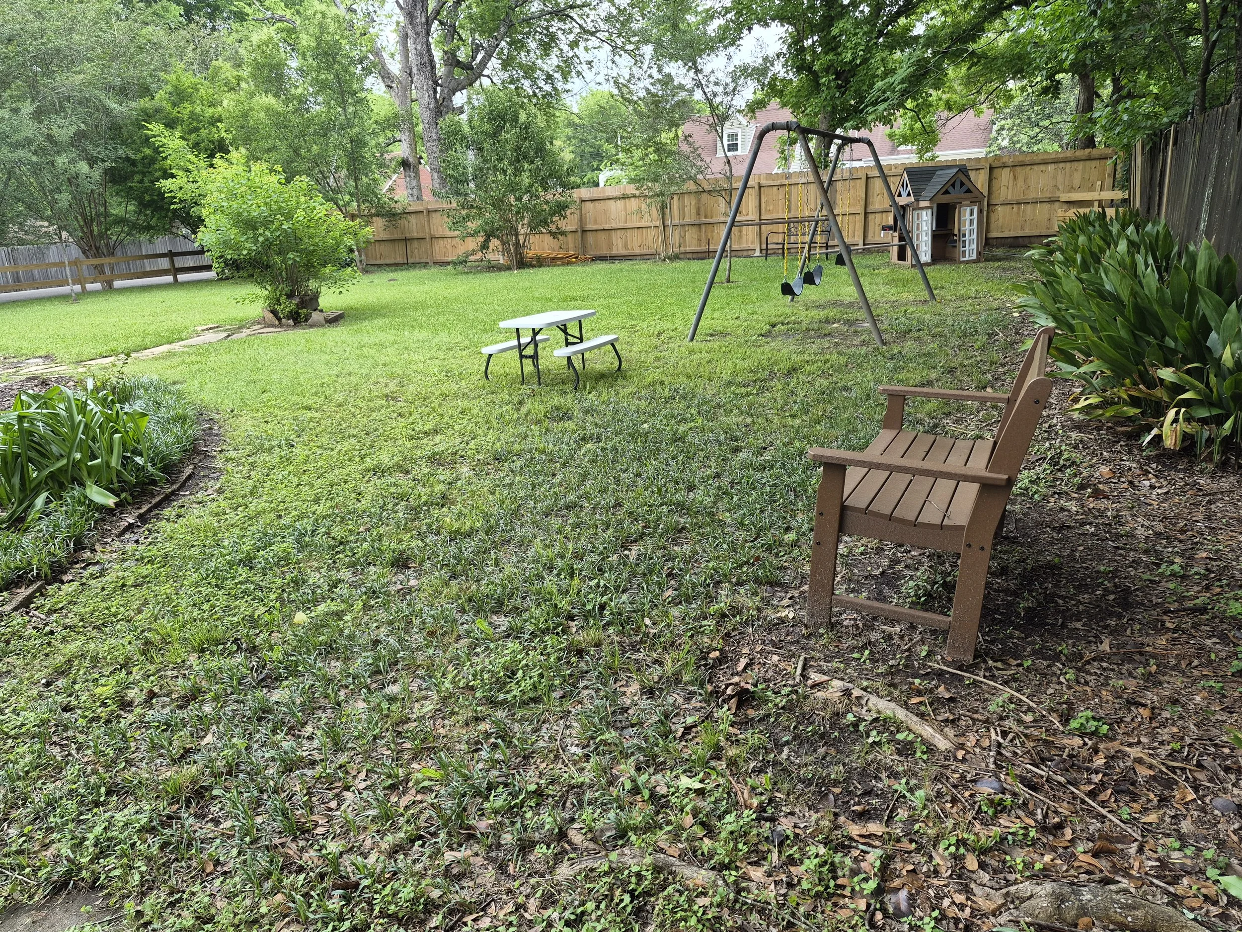 A backyard with green grass, trees, a wooden fence, a small playground with swings, a children's playhouse, and a garden bed with green plants.