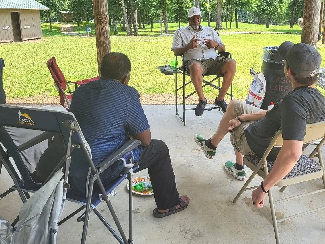Four men are sitting on folding chairs under a pavilion in a park, casually conversing. One man in a white shirt and hat is speaking, while the others listen. There are trees and a grassy area in the background.