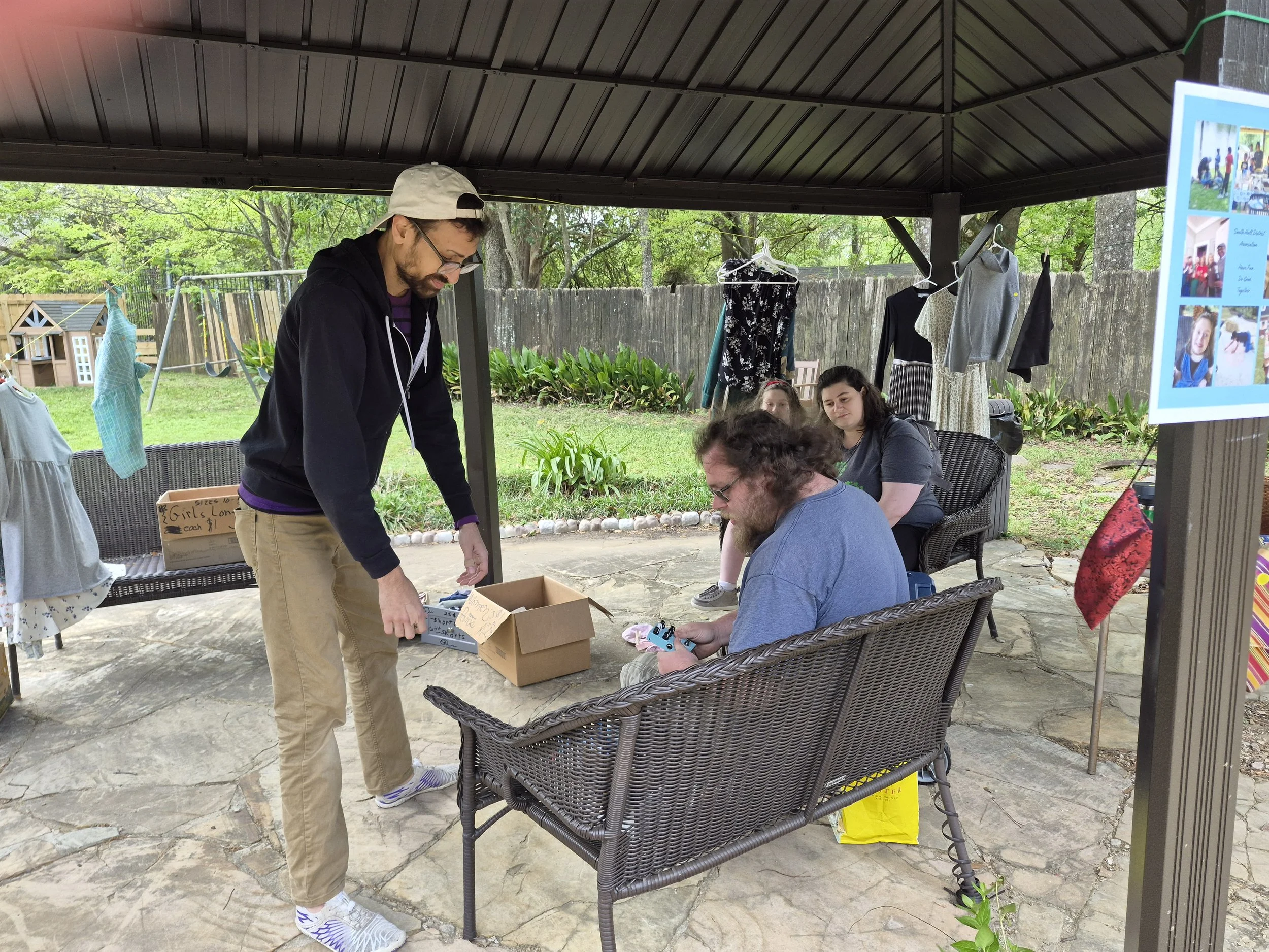 A group of people sitting and standing under a patio that has a dark roof. There are chairs, a table, and hanging clothes on a clothesline. One person is standing with glasses and a beige cap, and there are others seated, one of whom is holding a phone. A wooden box with signs and a pile of yellow bags are visible.