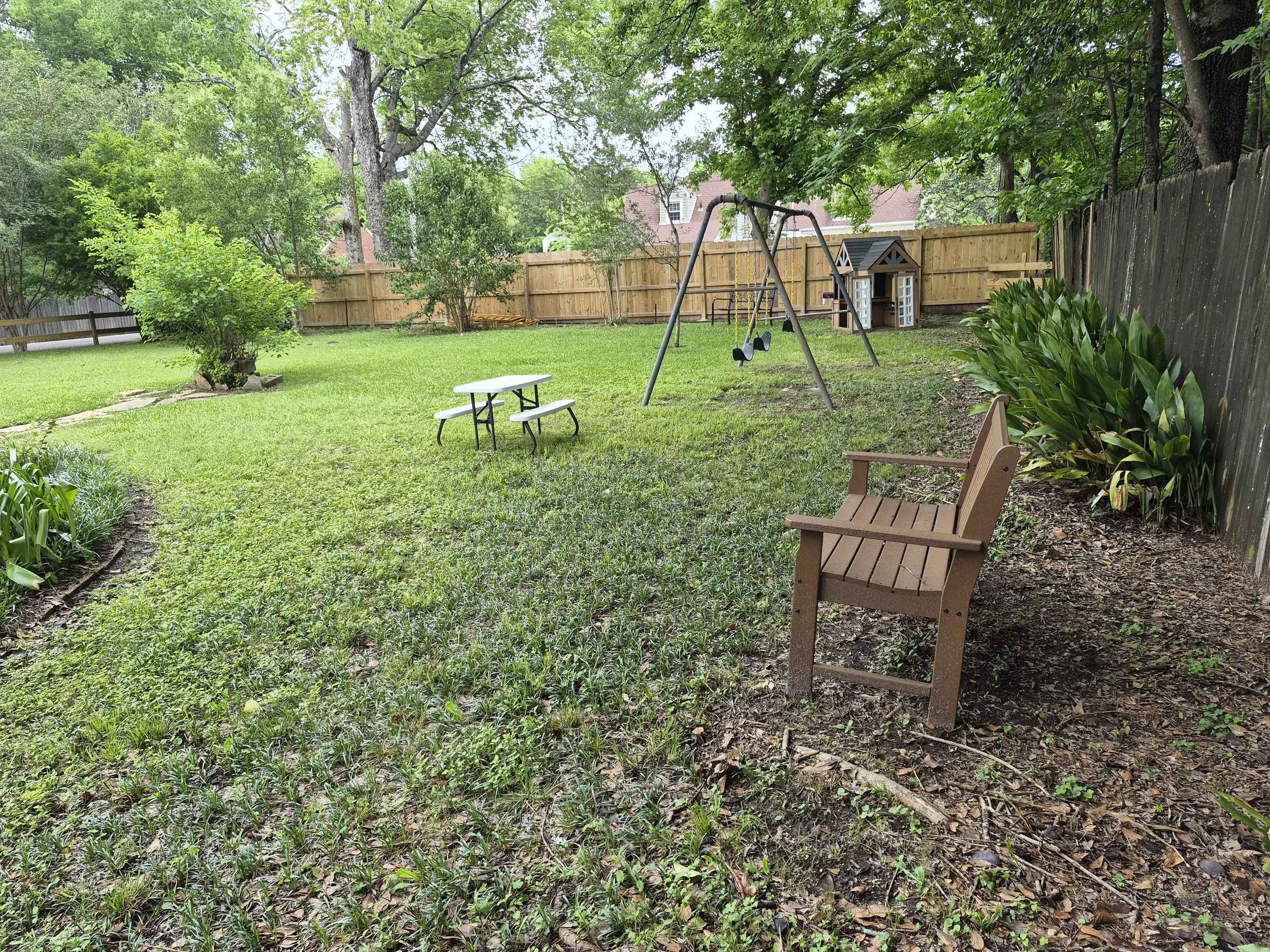 A backyard with green grass, trees, a wooden fence, a small playhouse, and a sitting bench, with a swing set and gardening plants.