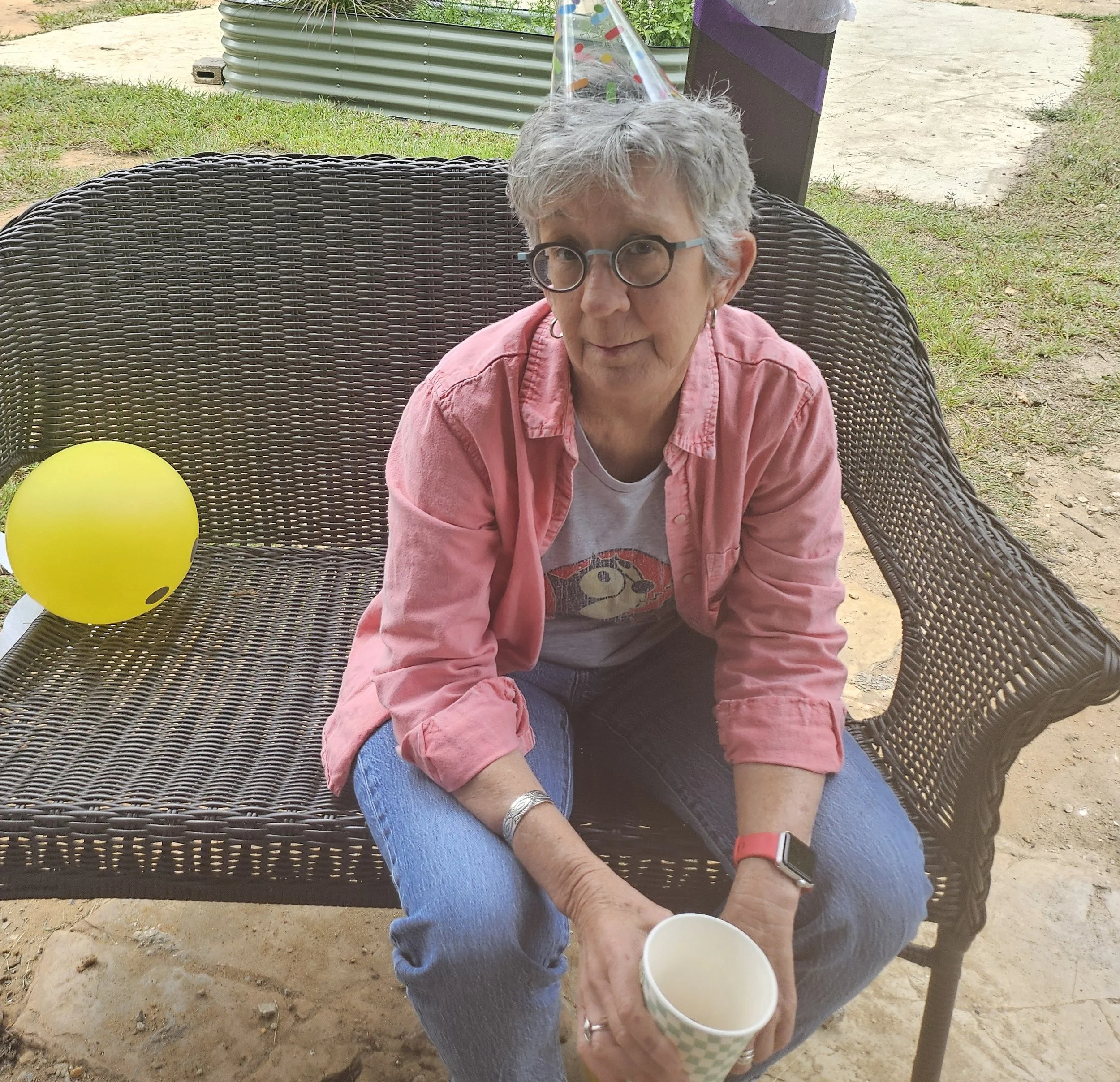 Older woman with gray hair and glasses sitting in a wicker chair outdoors holding two paper cups; a yellow balloon is visible on the table nearby, and a lick of green grass, concrete path, and gardening elements are in the background.