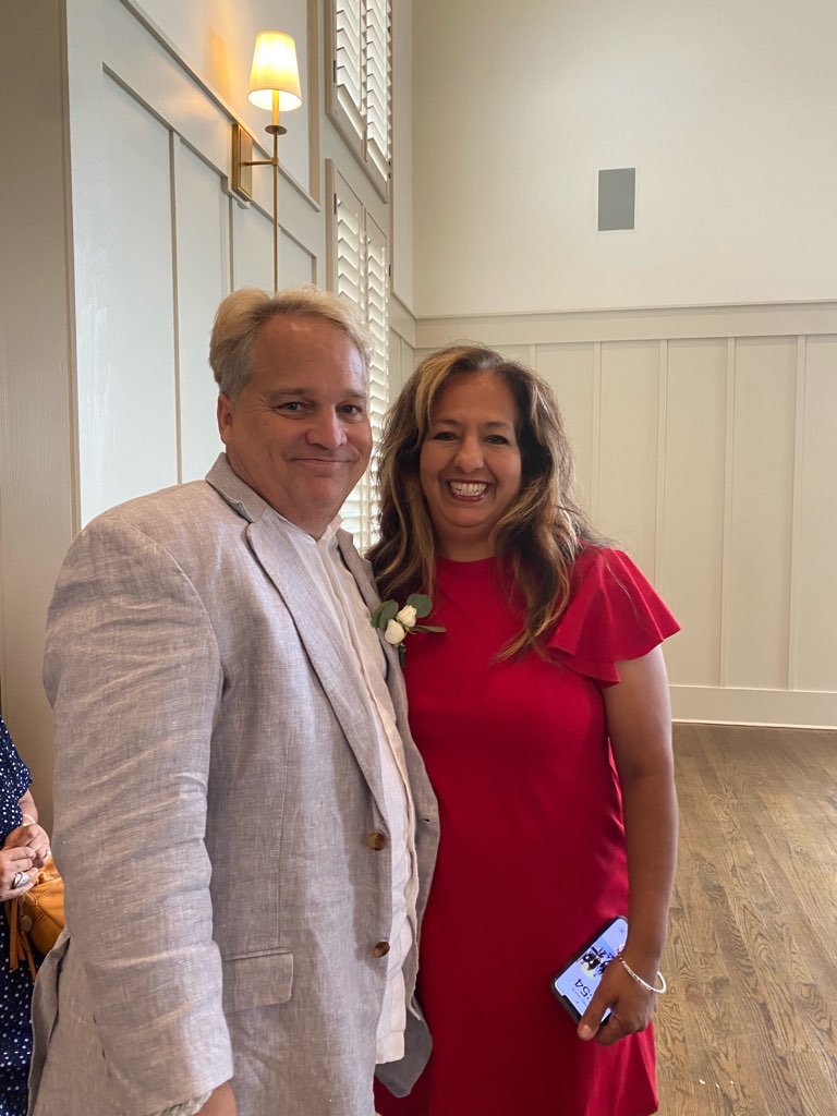 A man and woman smiling and posing for a photo at an indoor event, with the man in a light-colored linen suit and the woman in a red dress holding a phone.