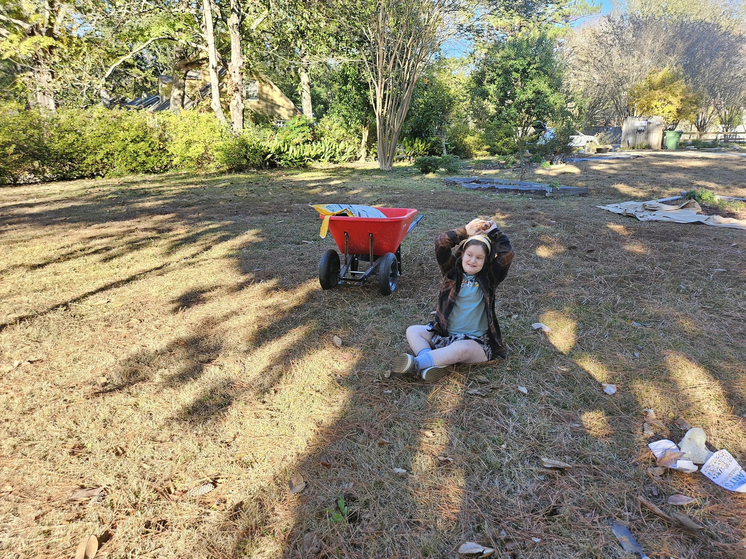 A young girl with curly hair is sitting on the grass in a yard, smiling. She is holding her head with both hands. There is a red wagon nearby with some packages inside. The yard has trees, bushes, and some scattered paper and debris. In the background, a house and a shed can be seen.