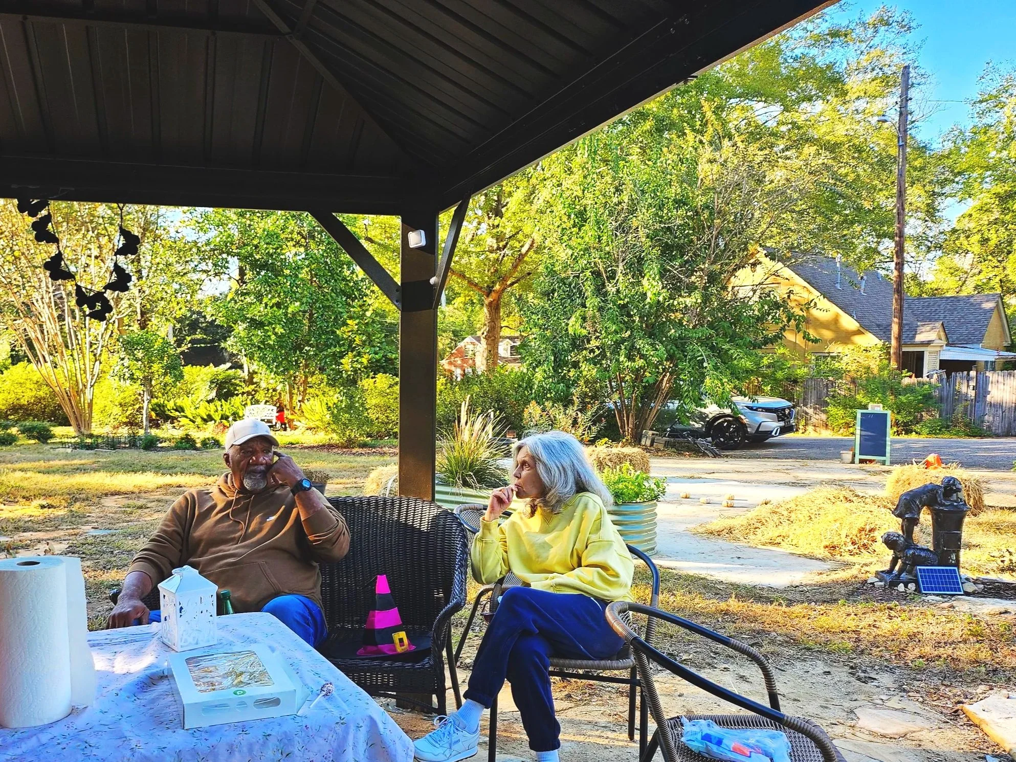 Two elderly people sitting outdoors under a roofed patio in a backyard, with trees, cars, and houses in the background. The man is wearing a brown shirt and white cap, and the woman is in a yellow jacket. They appear to be engaged in conversation.