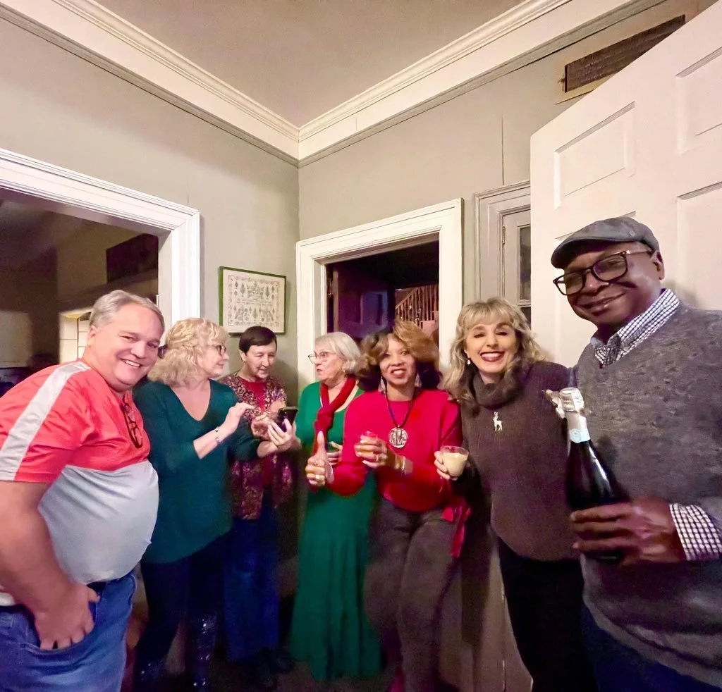 Group of people at a social gathering, smiling and holding drinks in a cozy indoor setting.