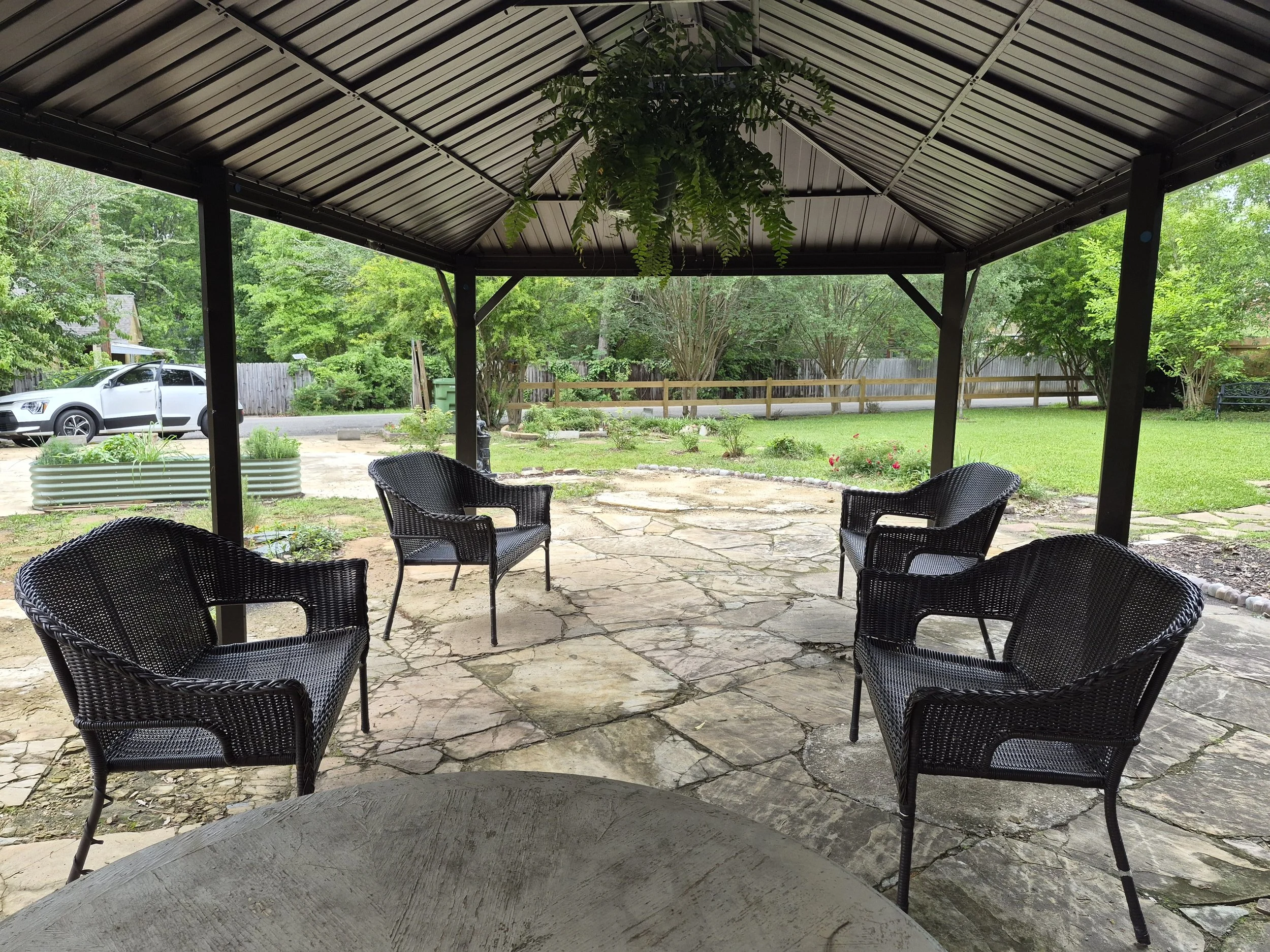 View from under a patio cover with black chairs around a stone patio, looking out onto a green backyard with trees, a wooden fence, and a parked car.