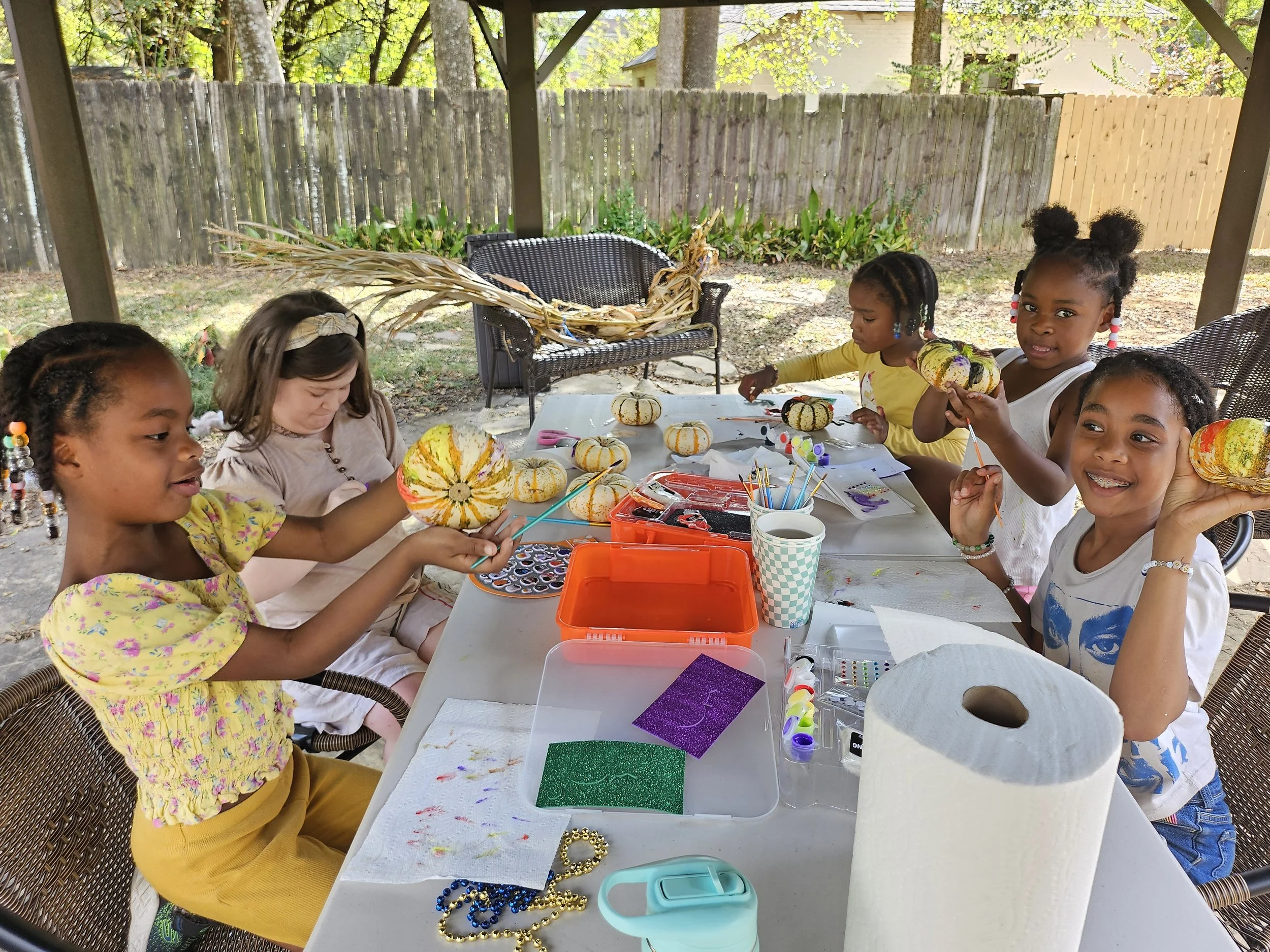 Group of young girls engaged in arts and crafts activity at an outdoor table under a covered patio.
