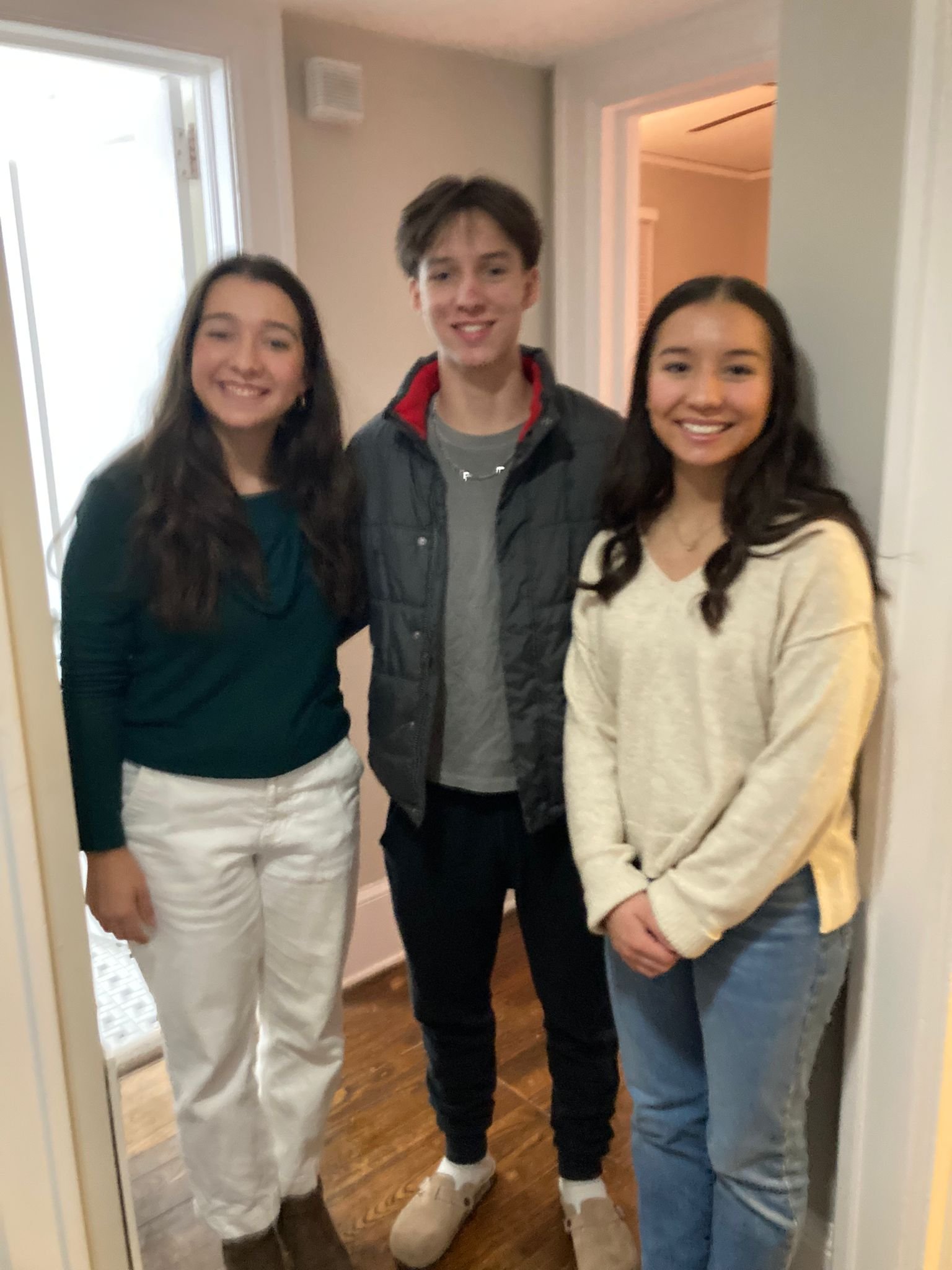 Three teenagers standing together indoors, smiling at the camera. The girl on the left is wearing a dark green top and white pants, the boy in the middle is wearing a dark jacket over a gray shirt, and the girl on the right is wearing a cream-colored sweater and blue jeans.