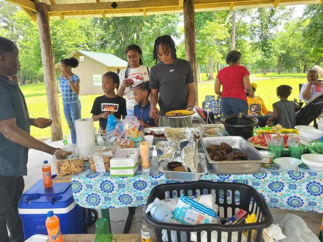 A group of people at a picnic table outdoors, with food and snacks, under a shelter with trees in the background.