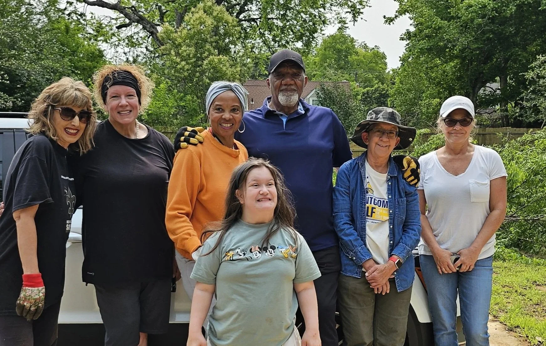 A group of seven people, including six women and one young girl, standing outdoors in a backyard with trees in the background. They are smiling and appear to be enjoying a gathering.
