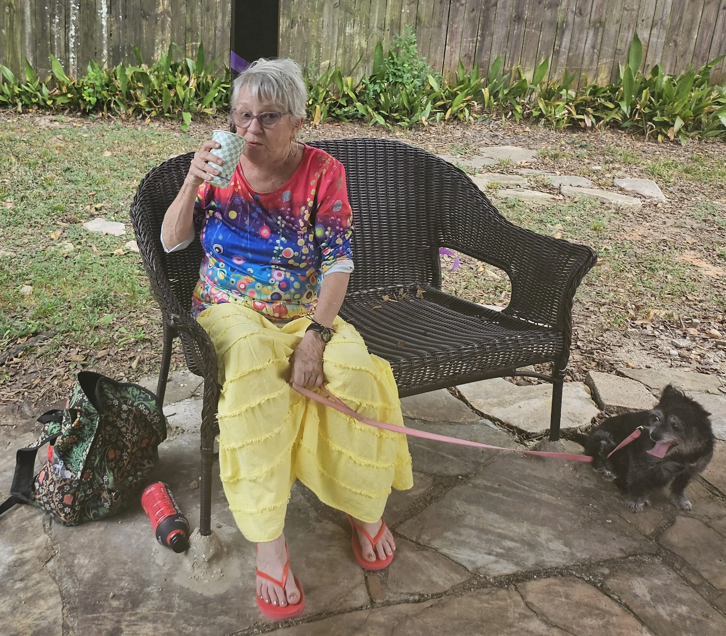 Older woman with short gray hair, glasses, wearing a colorful top and yellow skirt, sitting on a wicker chair outdoors, drinking from a checkered cup, with a black cat at her feet, surrounded by greenery and a wooden fence.