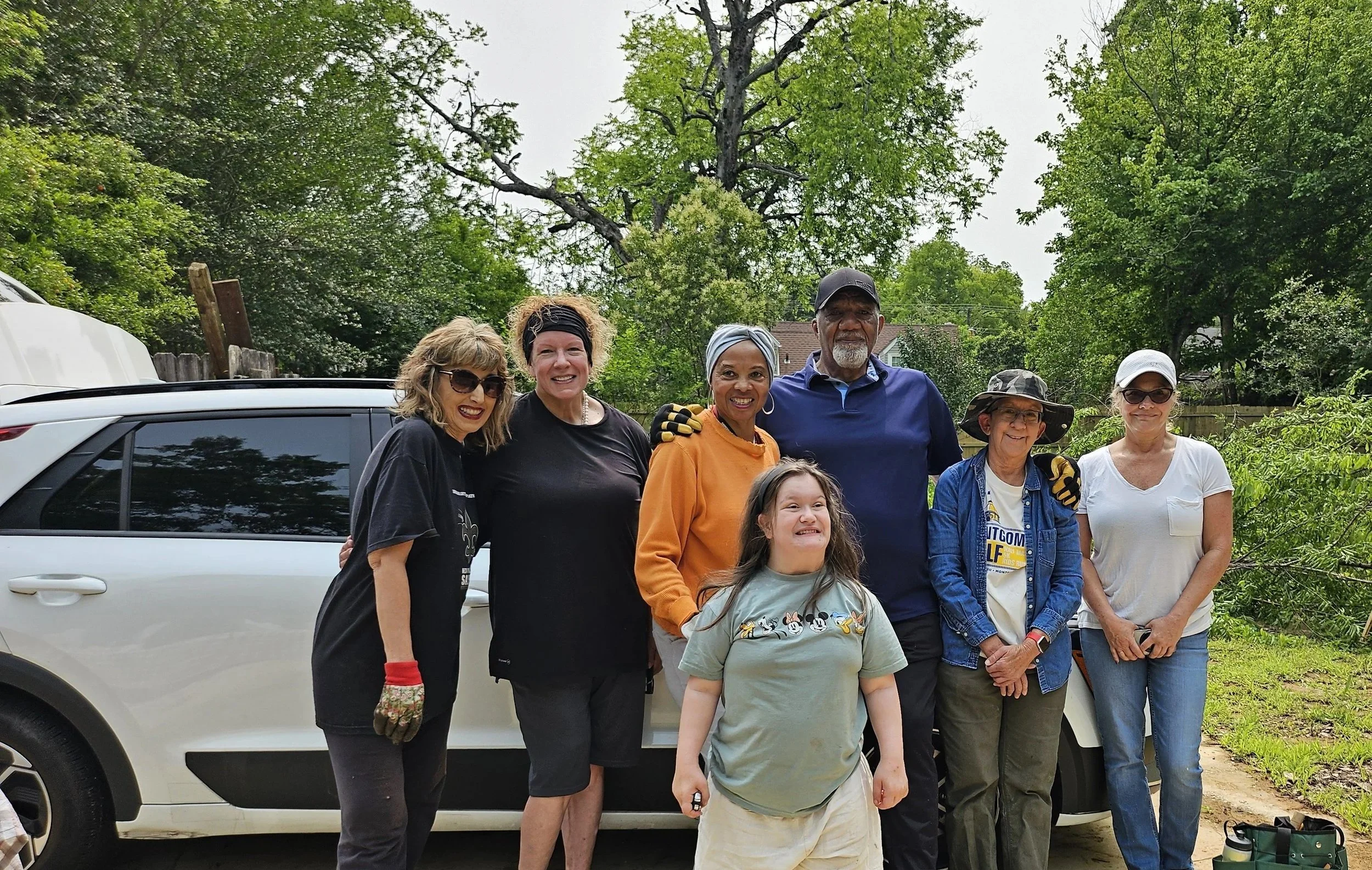 A group of seven people, including children and adults, standing outdoors in front of a white SUV and greenery, smiling at the camera on a cloudy day.