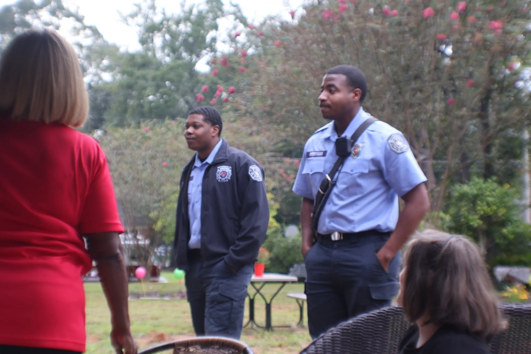 Two police officers and a woman having a conversation outdoors in a park with trees and pink flowers, during daytime.