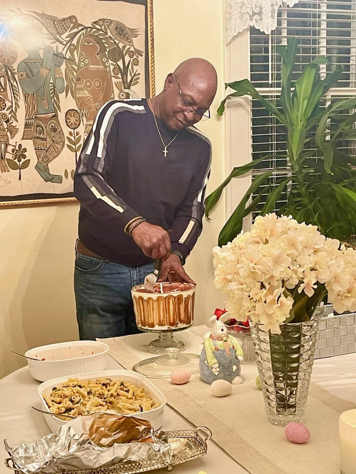 A man is cutting into a cake at a celebration dinner. The table has food, flowers, and decorative items, including a bunny figurine and colorful Easter eggs.