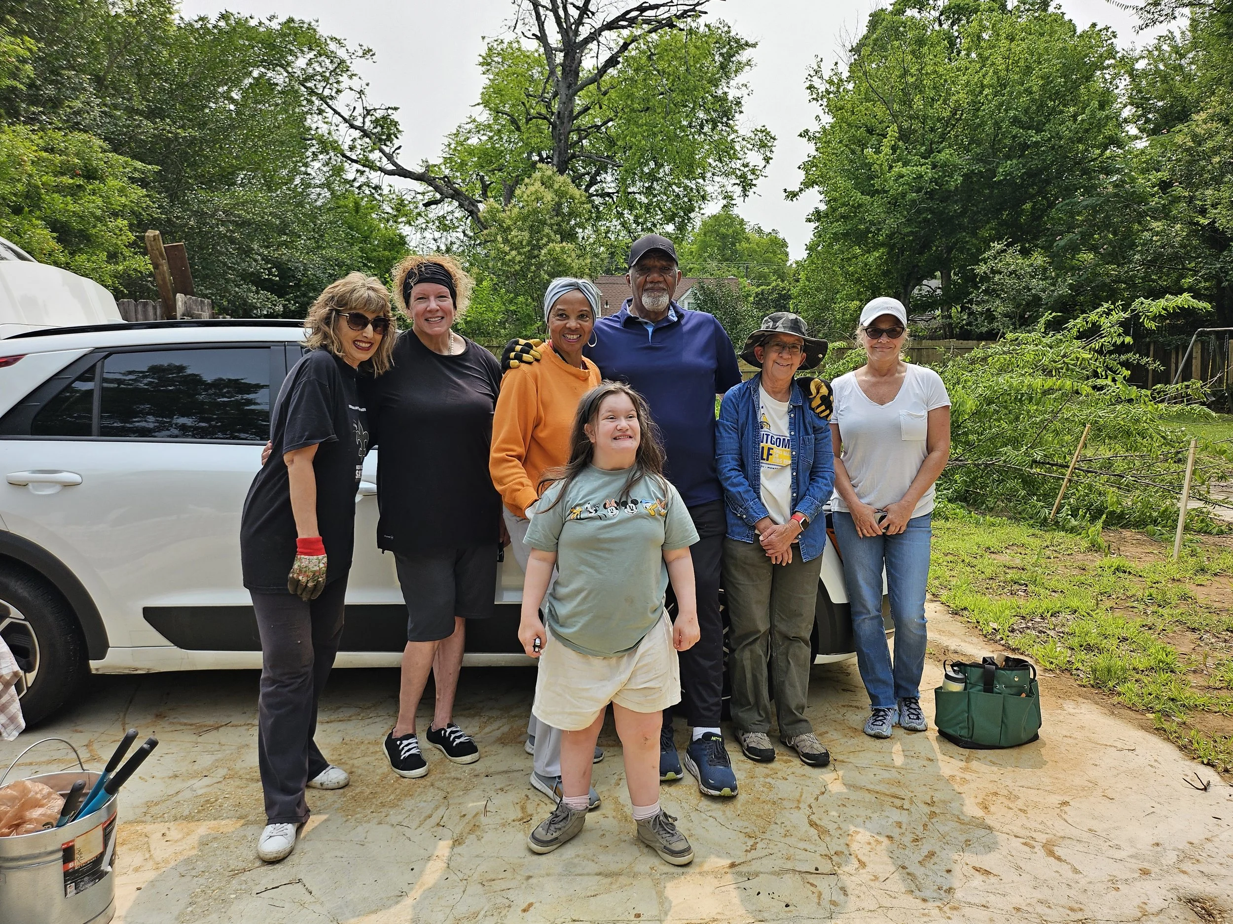 A group of seven diverse people standing together outdoors in a backyard with green trees and a wooden fence in the background, smiling for the photo.