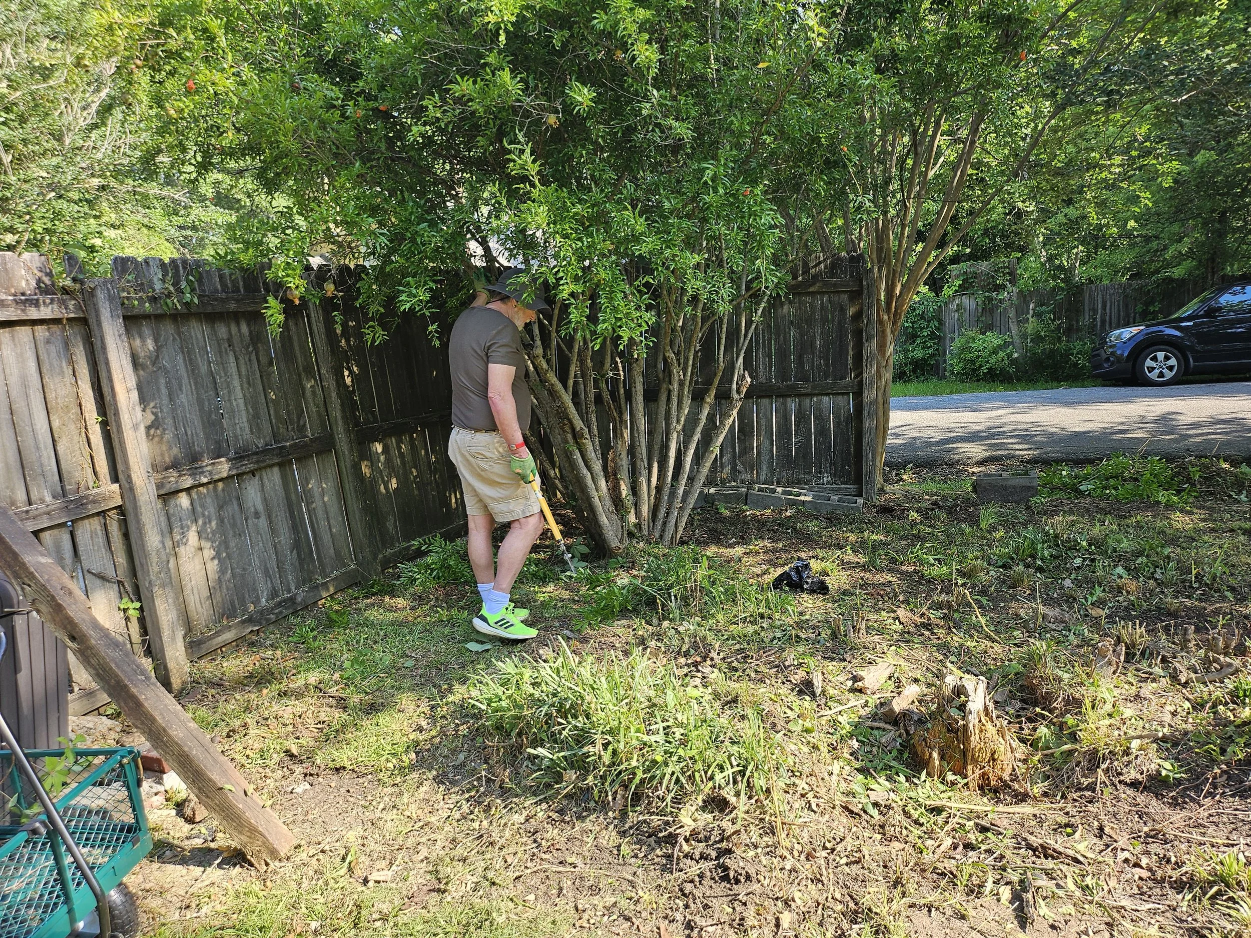 A man wearing a black hat, black shirt, beige shorts, and bright neon green sneakers is gardening or yard work in a backyard. He is standing near a black wooden fence with a large leafy green tree beside him. There are various plants, some gardening tools, and a parked black car in the background.