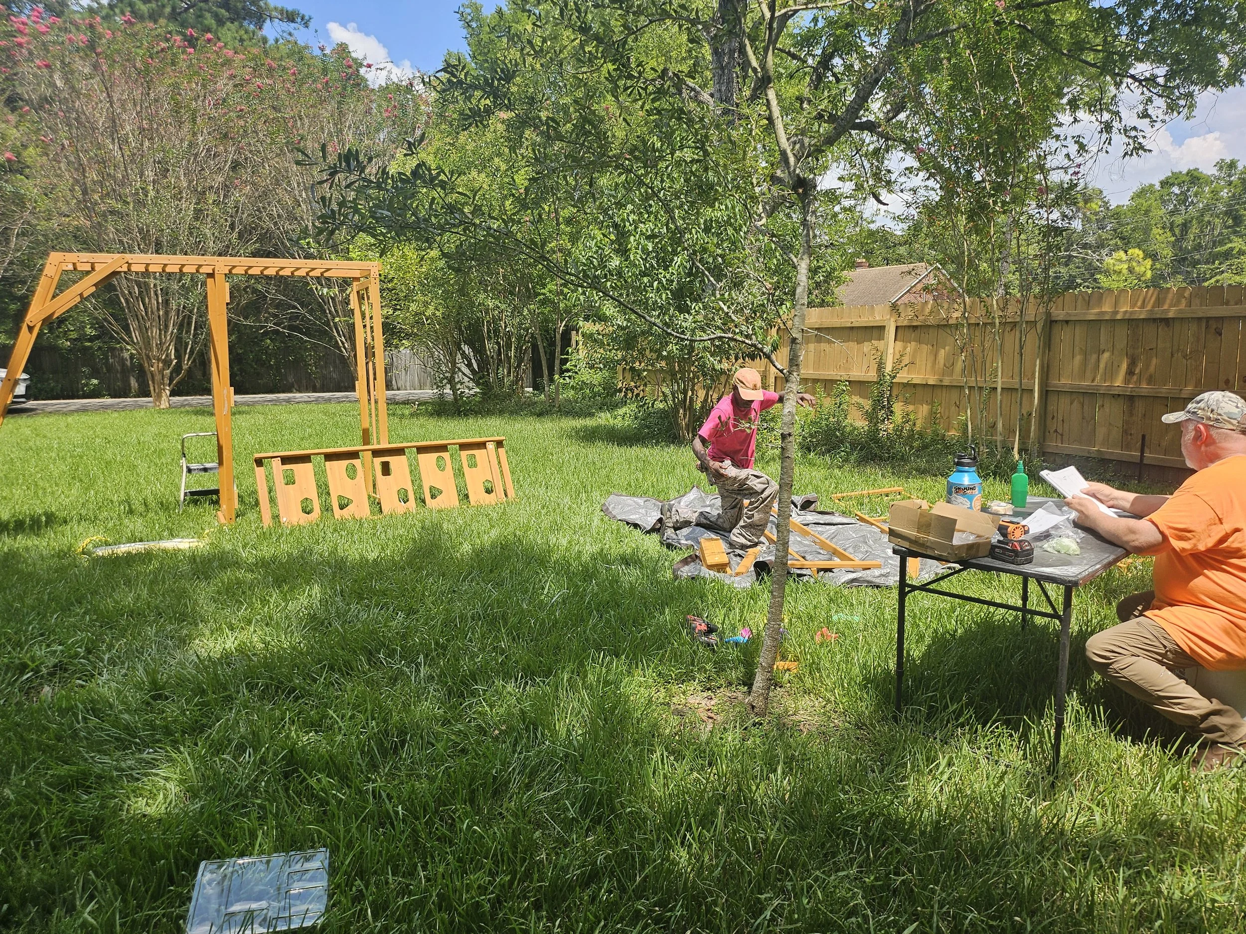 Two people working on planting a small tree outdoors in a backyard during the daytime. There is gardening equipment and a table with supplies nearby, a wooden fence, and trees in the background.