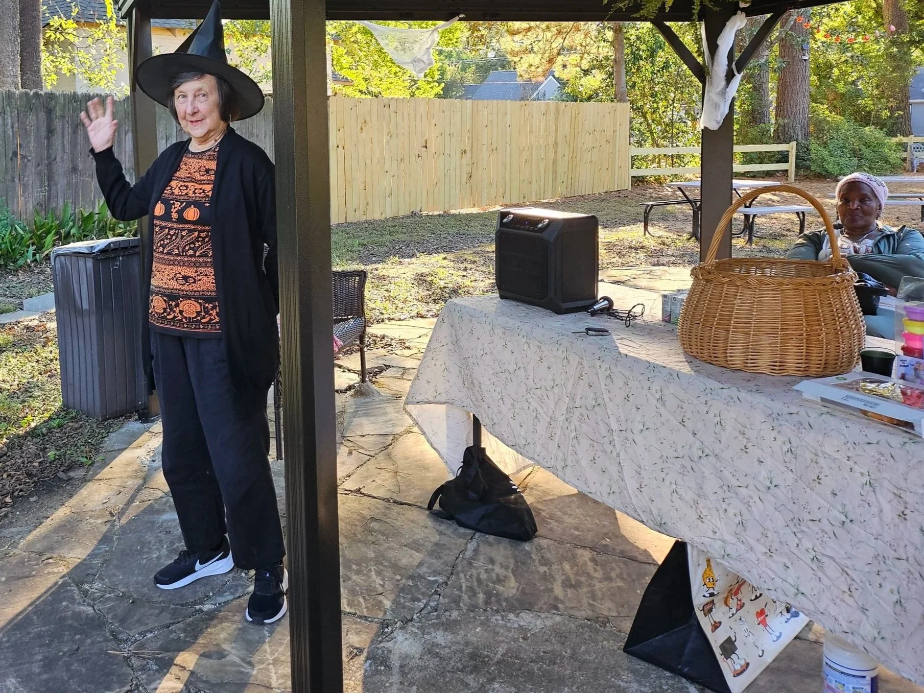 An elderly woman dressed as a witch, wearing a black hat and Halloween-themed shirt, standing outside near a table with a white tablecloth. A woman is sitting at the table, and Halloween decorations, including a skull prop and a hanging ghost, are visible in the background.