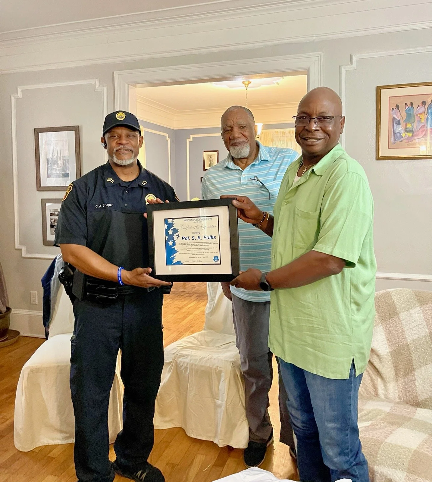 Three men standing indoors, two of them holding a framed certificate. The man on the left is in a police uniform, the man in the middle is wearing a light blue striped shirt, and the man on the right is wearing a light green shirt. They are smiling, and the room has framed artwork and a door leading to another room in the background.