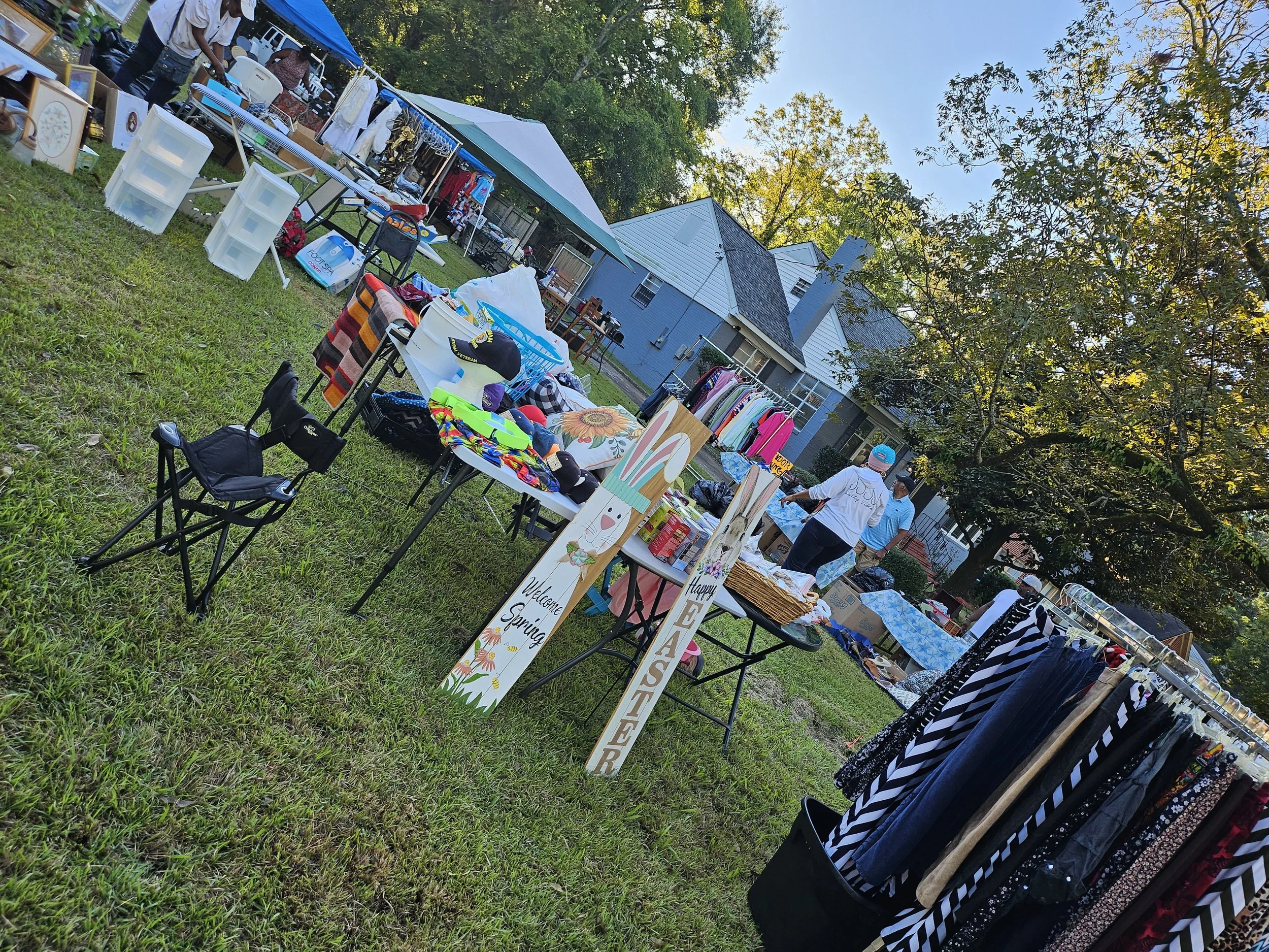 Outdoor yard sale in a backyard with tables displaying various items including clothes, signs, and decorative pieces. People browsing, trees with autumn foliage, and houses in the background.