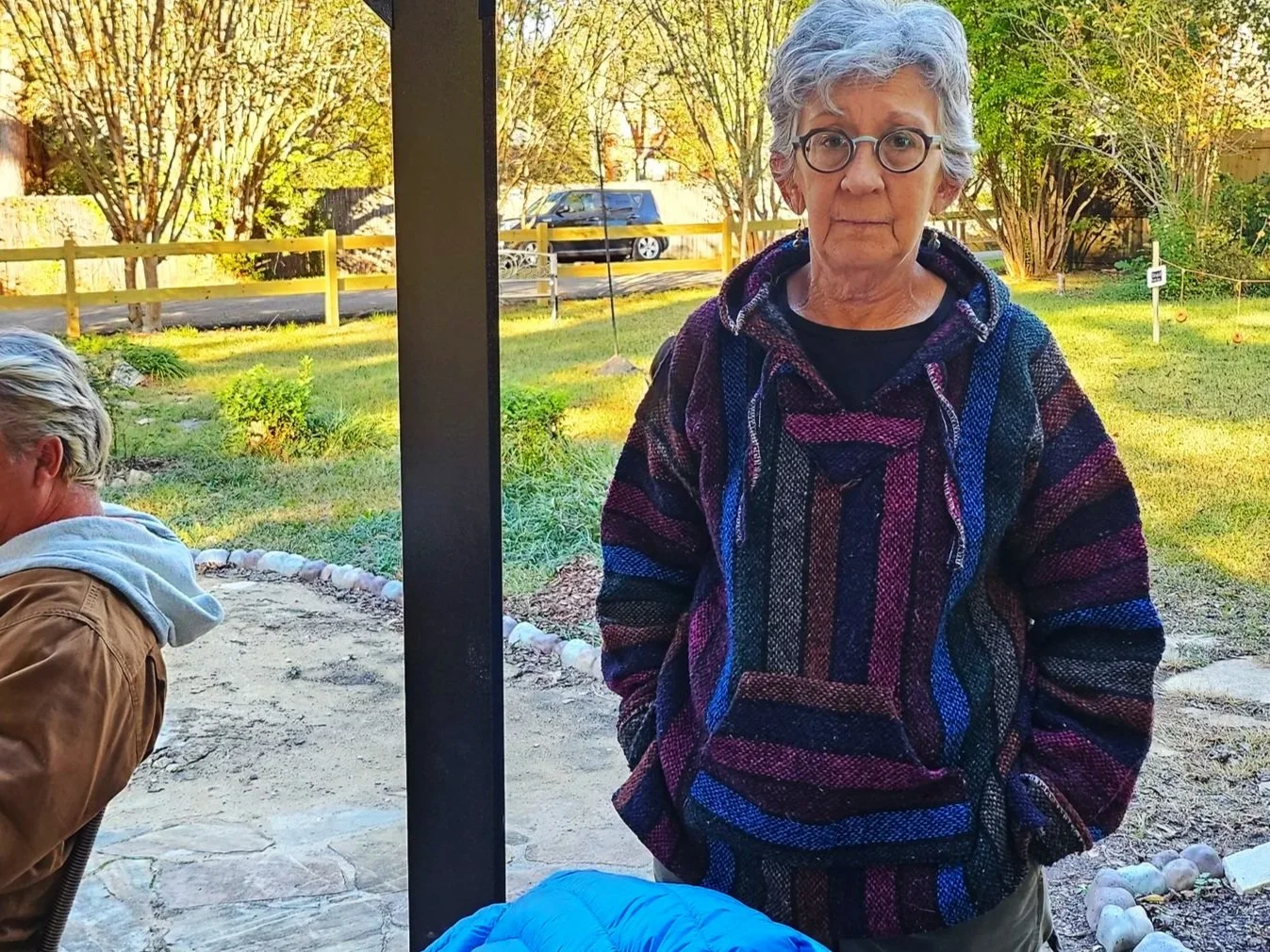 An older woman with gray hair and glasses wearing a colorful striped hoodie standing outdoors in a yard with trees, grass, and a wooden fence.