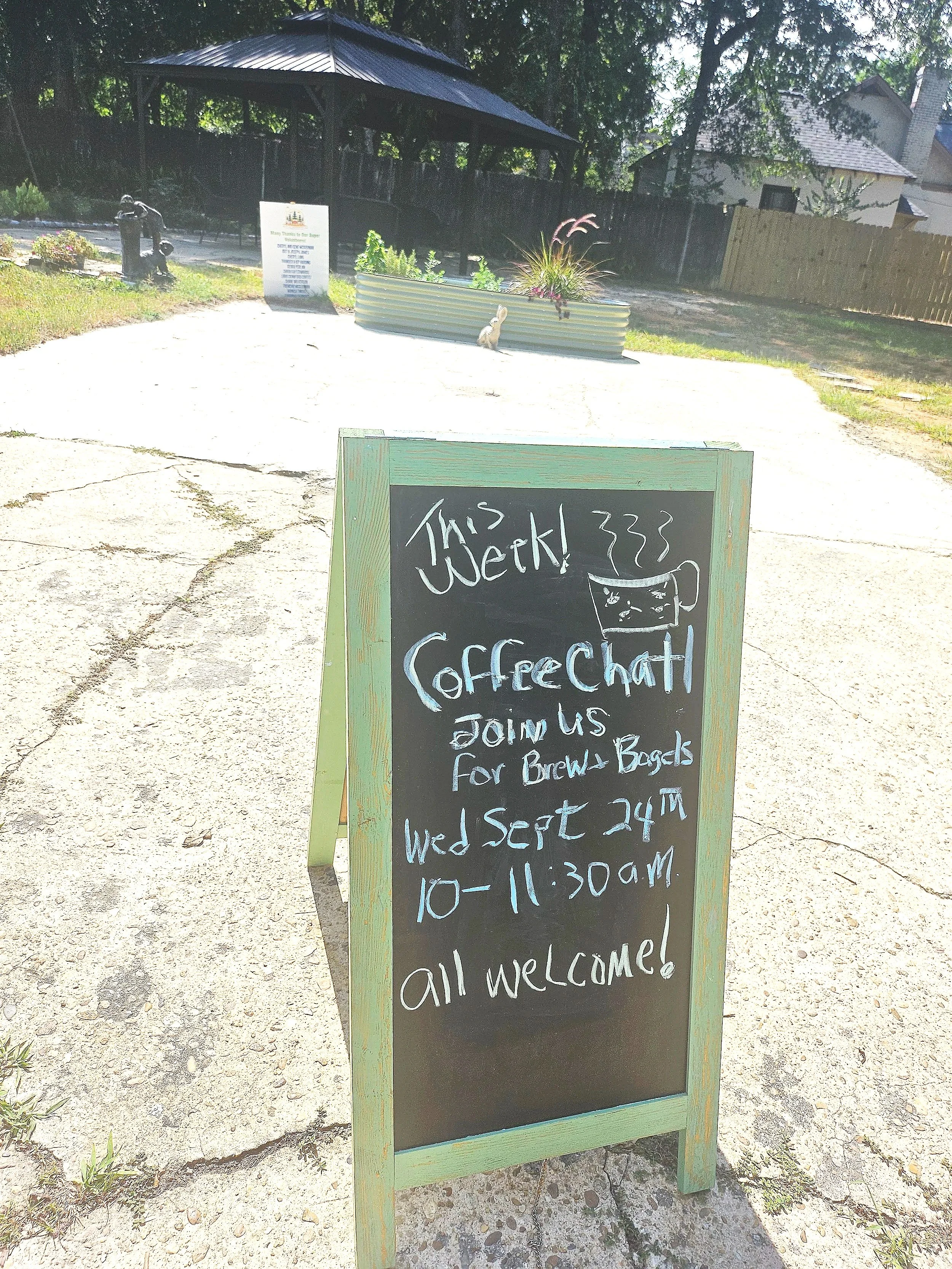 A chalkboard sign outside a cafe or coffee shop with handwritten text indicating the shop's hours and welcoming customers. In the background, there is a planter with plants, a shaded pavilion, and a fenced yard with trees and houses.