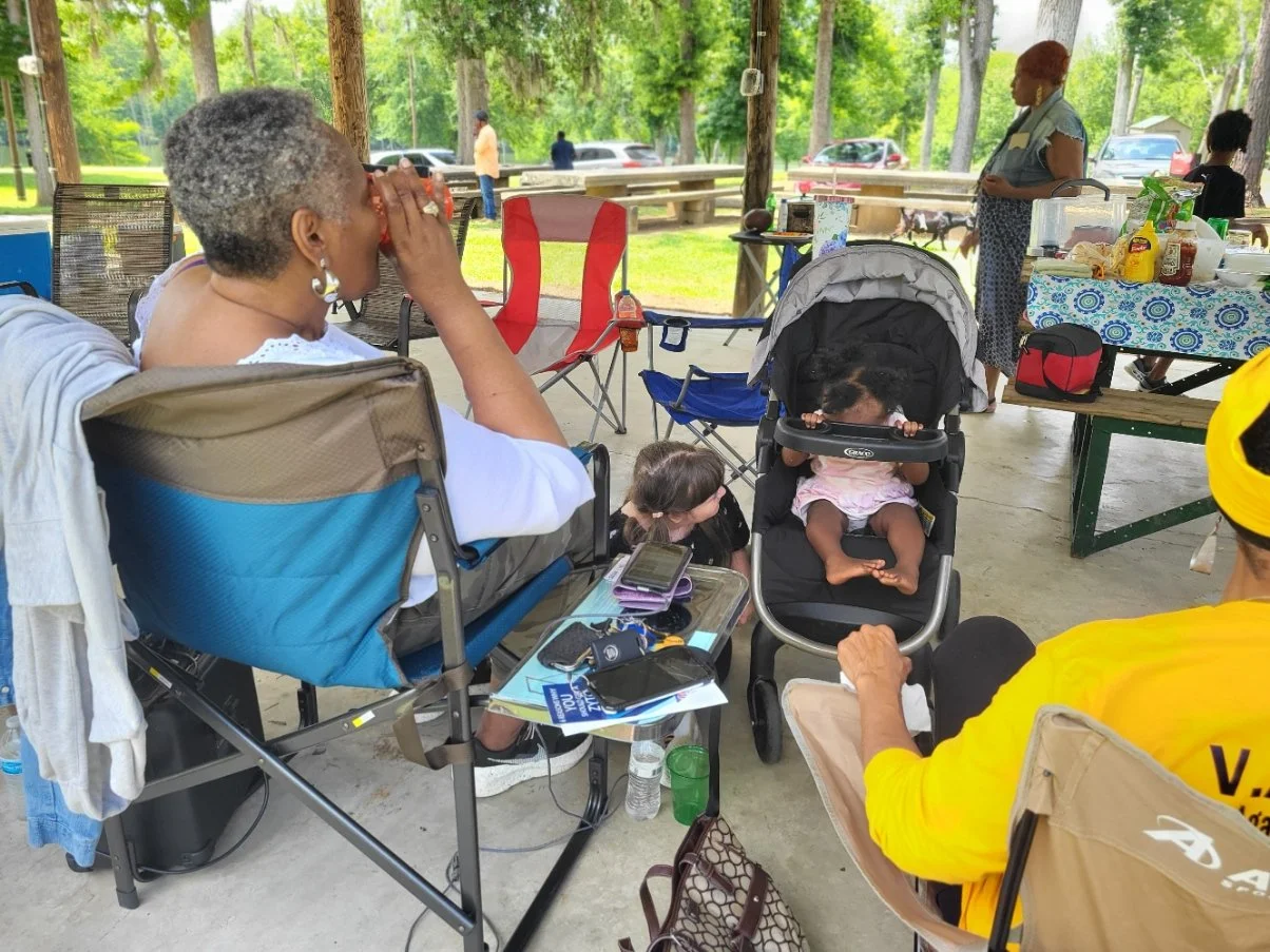 A group of people gathered at a daytime outdoor picnic under a pavilion with trees in the background. There are folding chairs, a table with food, drinks, and supplies, and children sitting in strollers.