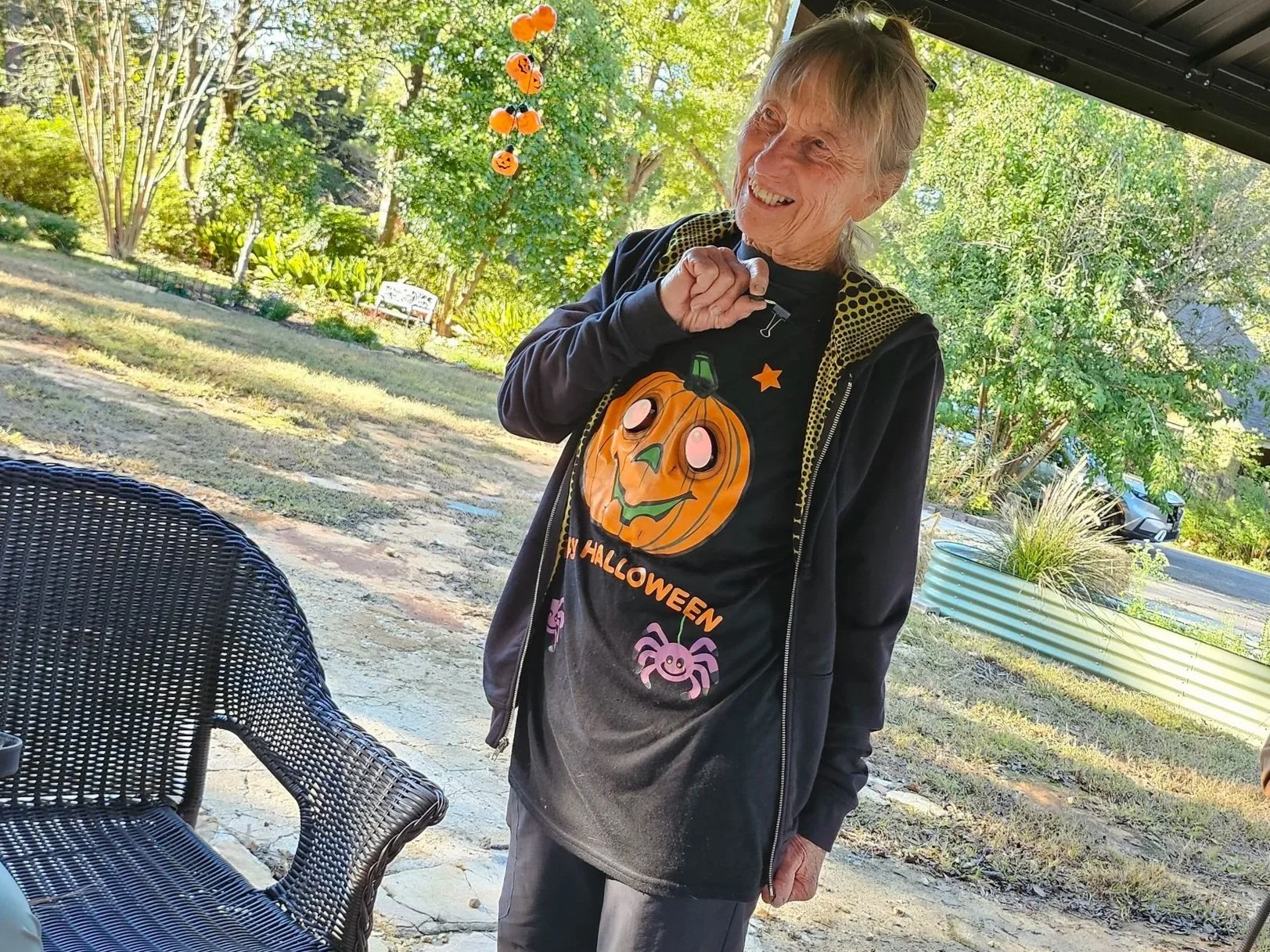 An elderly woman smiling outdoors in a backyard, wearing a black Halloween-themed shirt with a pumpkin face, spider and moon designs, and a black jacket, with Halloween decorations hanging in the background.