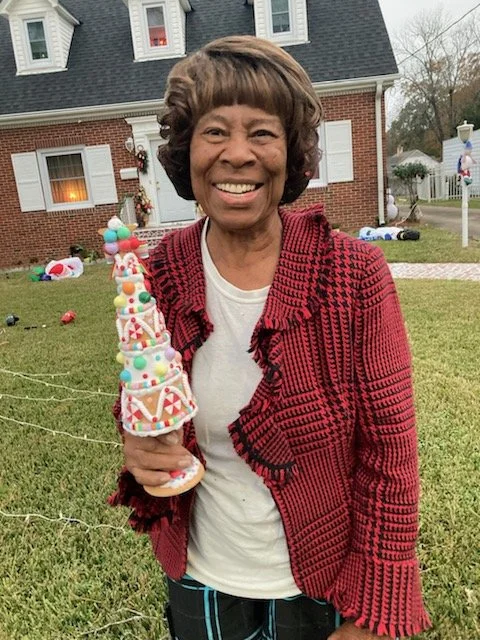 A smiling woman holding a decorated Christmas tree-shaped cone outdoors in front of a brick house.