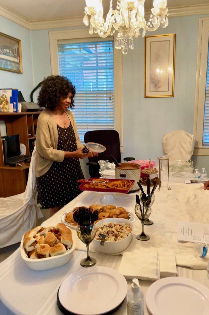 A woman standing by a dining table with food, holding a container and a spoon. The table has various dishes including rolls, salad, and a main course in a crockpot. The room has windows with blinds, framed artwork, and a chandelier.