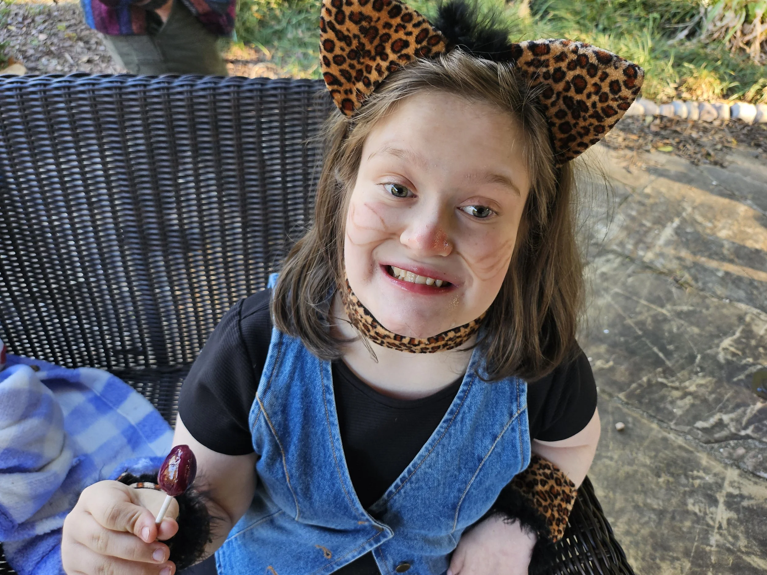 A young girl with light skin and long brown hair is sitting outdoors on a wicker chair, smiling at the camera. She is wearing a leopard print headband with matching ears, a black shirt, and a denim dress. She is holding a trunk-sized lollipop with a cherry at the top. The background includes greenery and a cobblestone path.