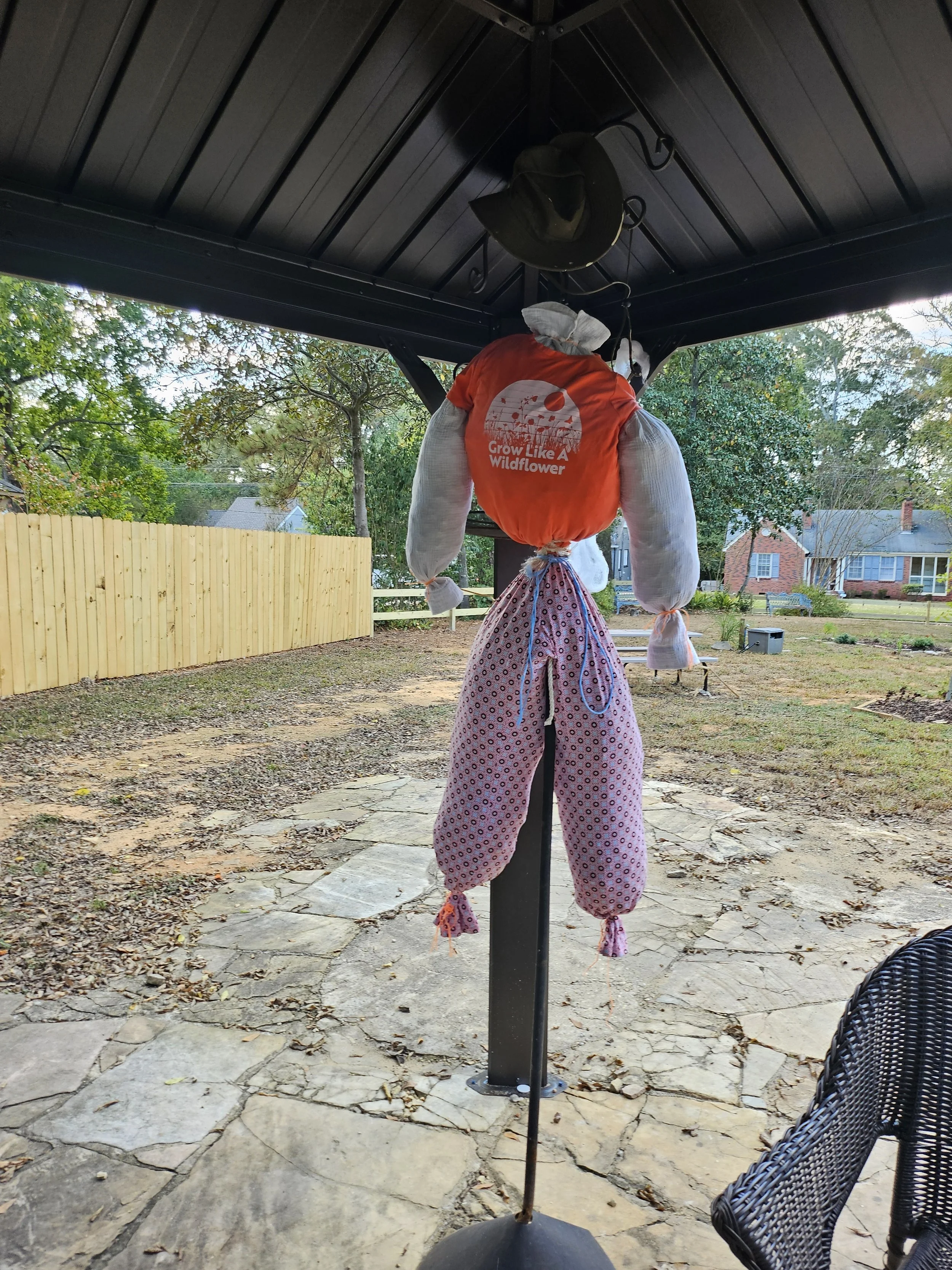 A scarecrow dressed in a red shirt with the words 'Grow Like A Wildflower' printed on it and pink patterned pants, hanging from a black pole under a porch roof.