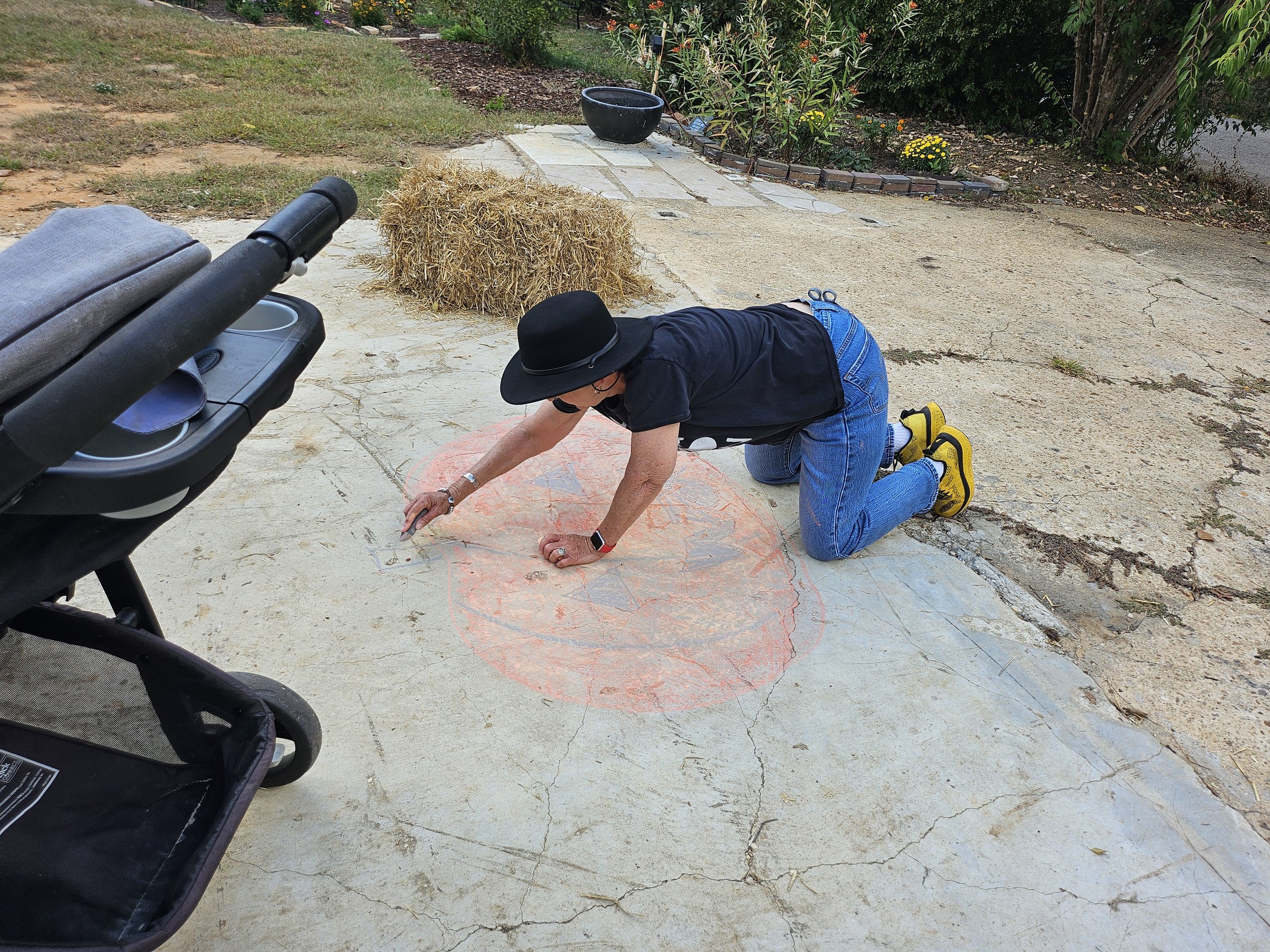 A person wearing a black hat, dark shirt, and blue jeans kneeling on concrete ground, drawing with chalk on the pavement. Nearby, a black stroller is visible in the foreground.